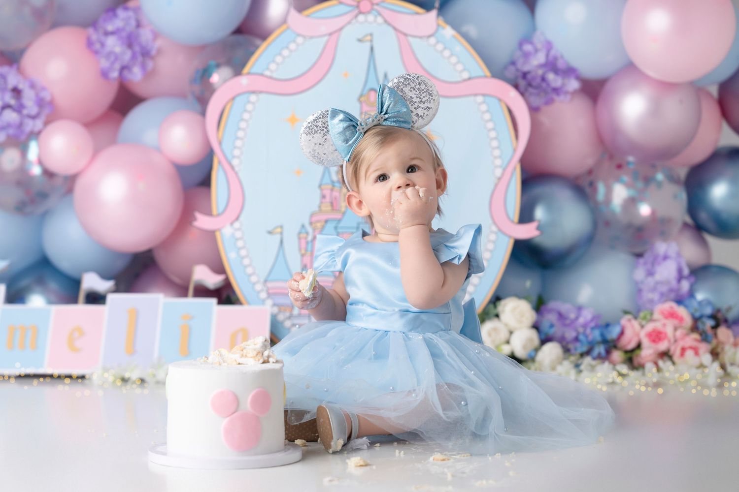Baby girl in blue tutu dress celebrates first birthday with cake smash, surrounded by pink and blue balloon backdrop.