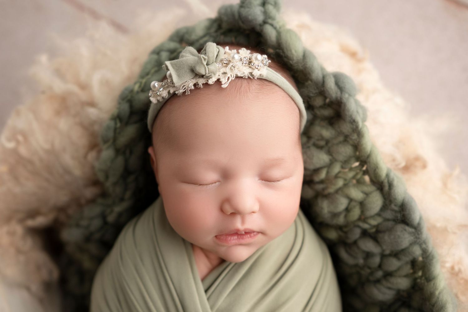 Sleeping newborn wrapped in sage green swaddle with delicate pearl headband on cream fur backdrop.