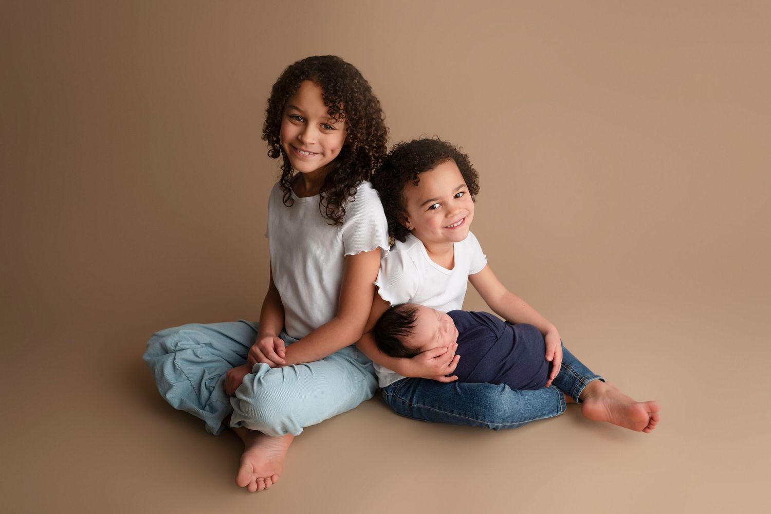 Two children in white tops and jeans sit together on a studio floor, smiling, with a newborn baby resting between them.
