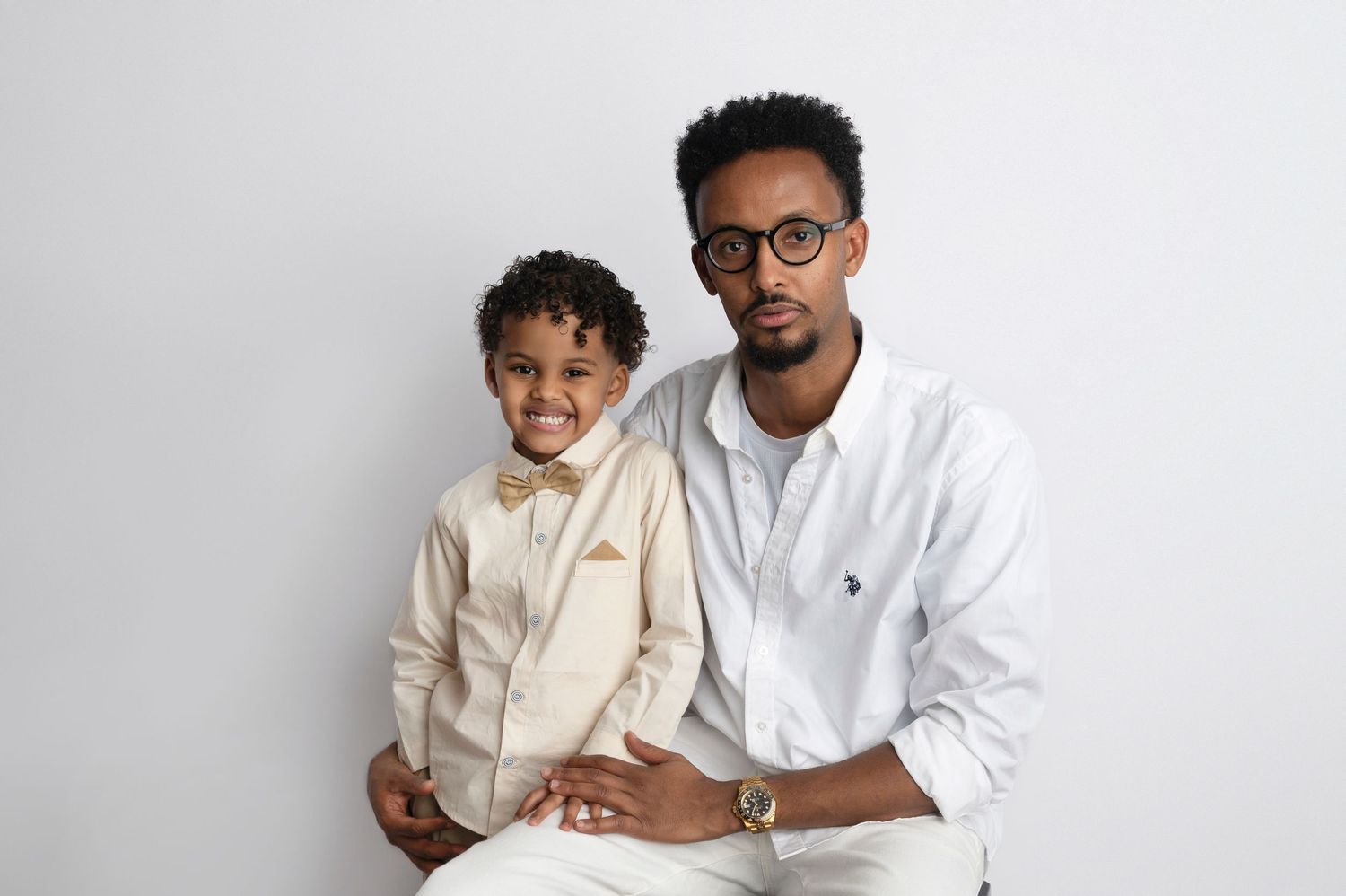 A man and young boy in matching white and beige outfits pose together against a white background.