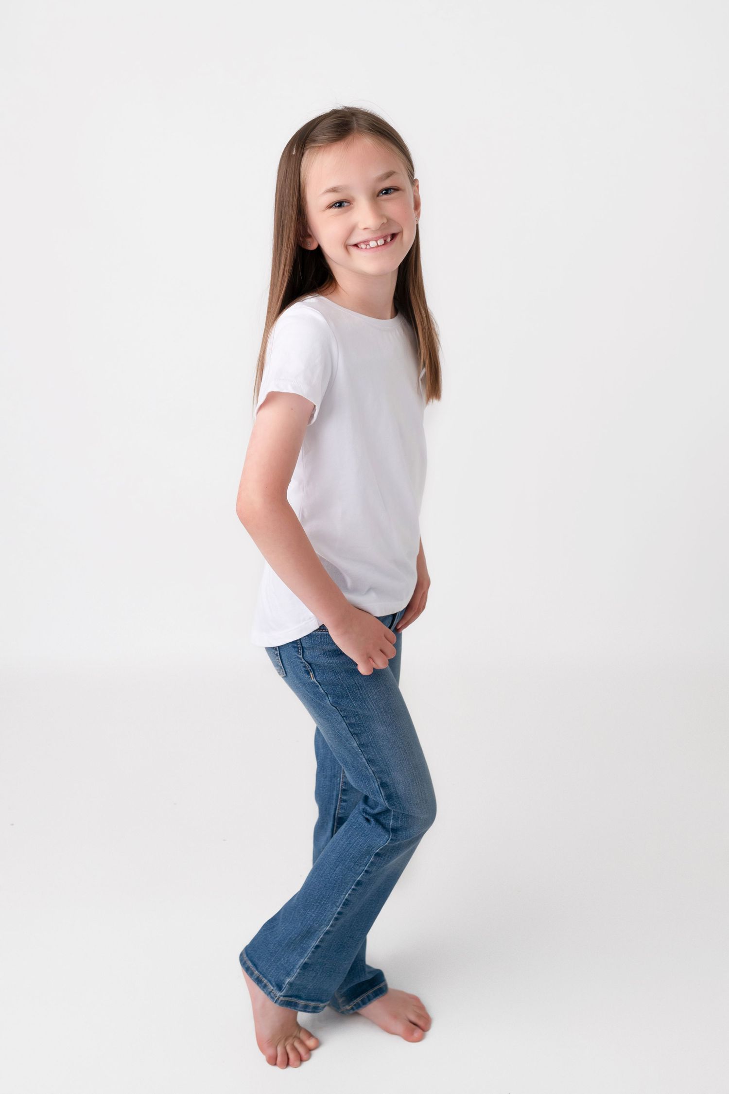 Smiling young girl in white t-shirt and blue jeans standing barefoot against a white background.