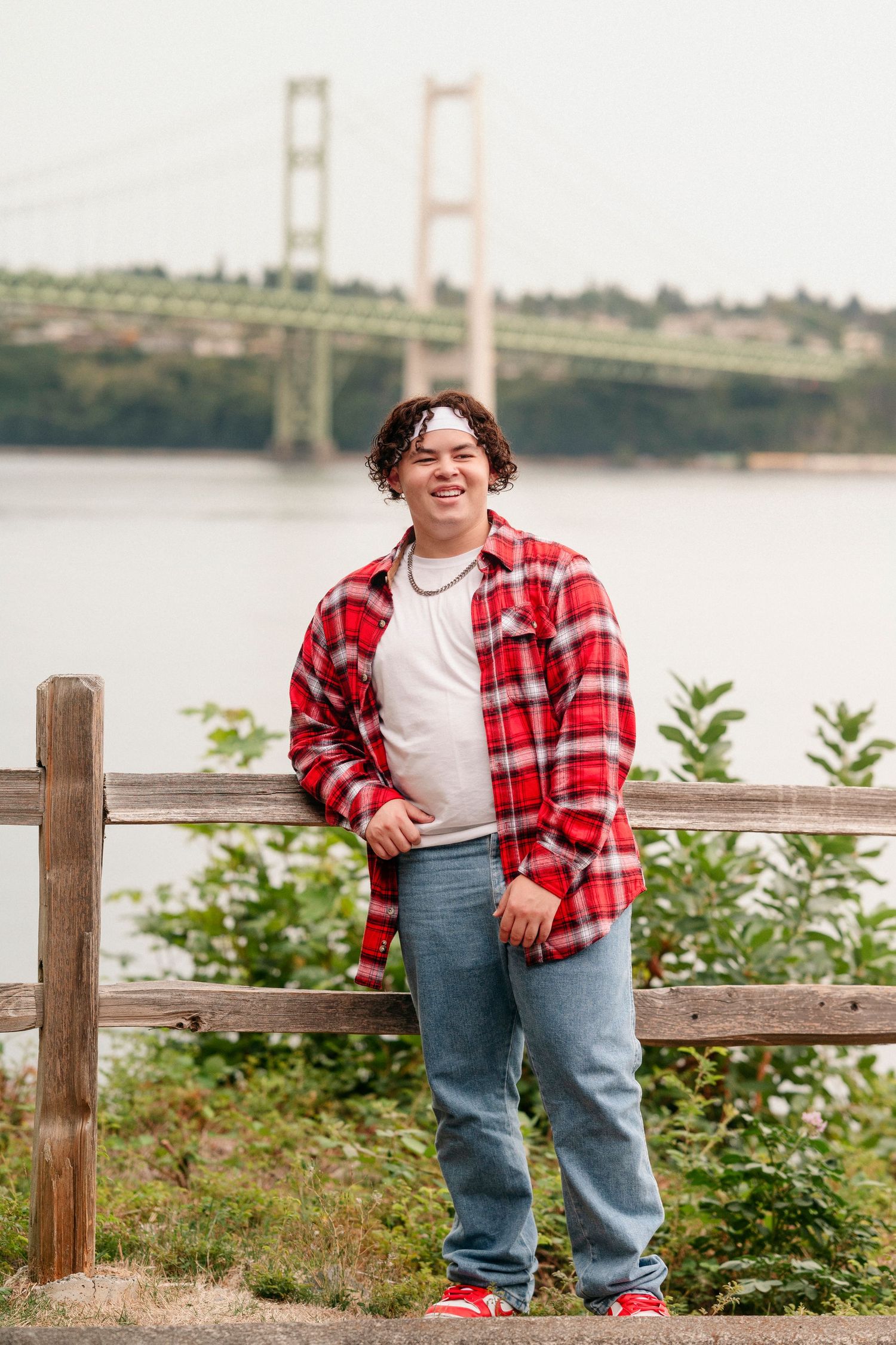 A person in a red plaid shirt and jeans poses near a wooden railing with water and trees in the background.