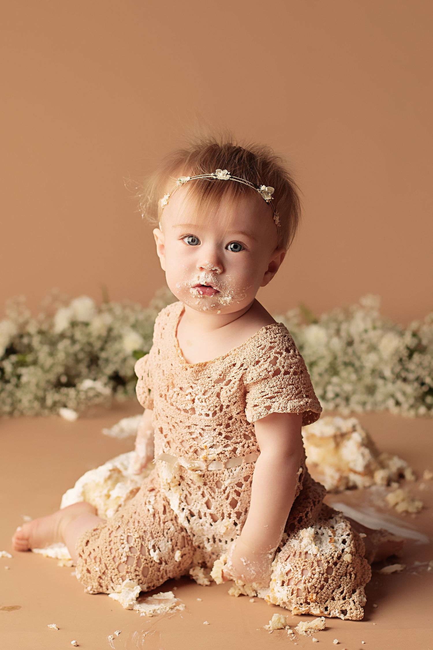 Sweet baby in lace dress sits among delicate white flowers during professional photoshoot with brown backdrop.