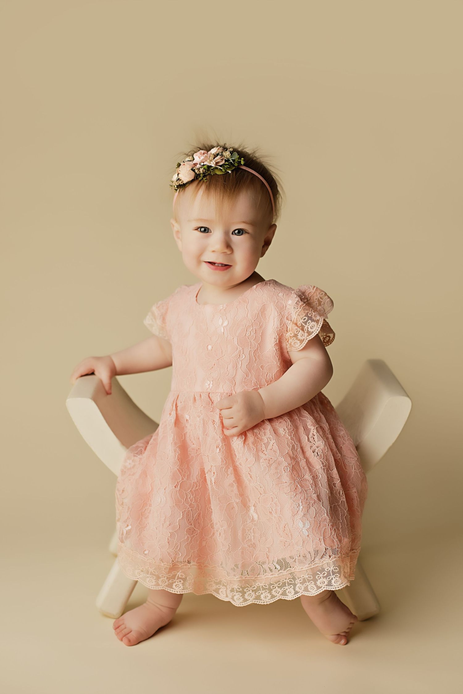 Sweet baby girl in peach lace dress poses during professional studio portrait session against beige backdrop.