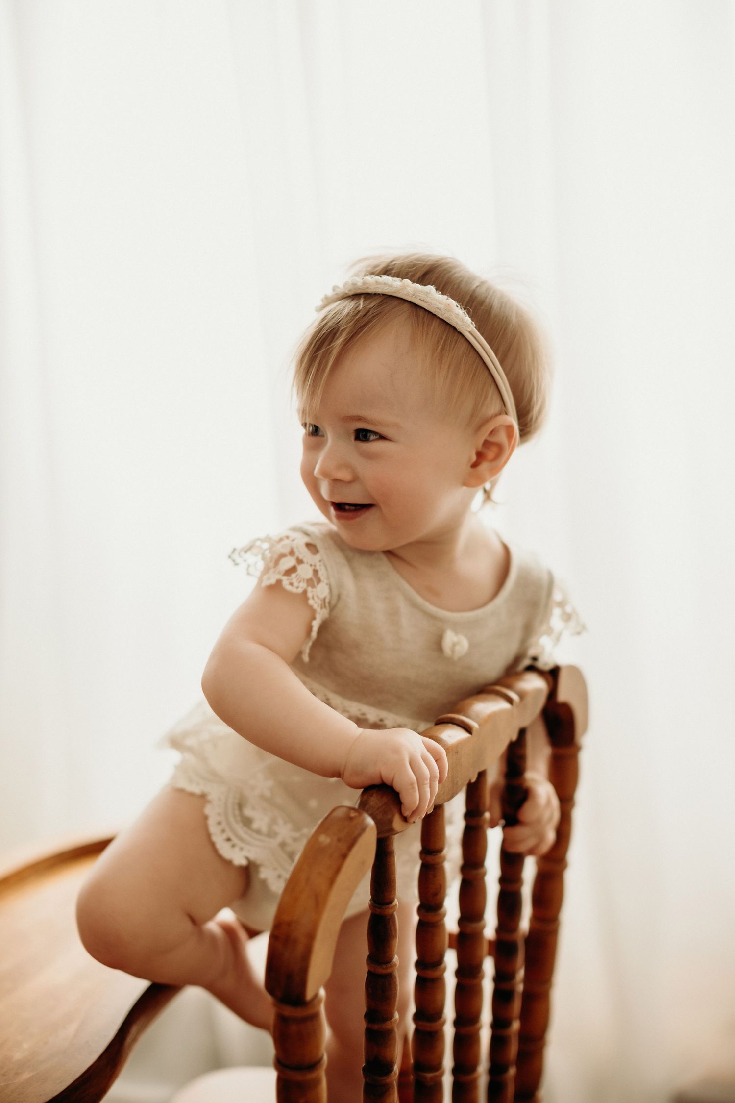 A happy baby in a cream dress leans and plays on a wooden chair in soft natural light.