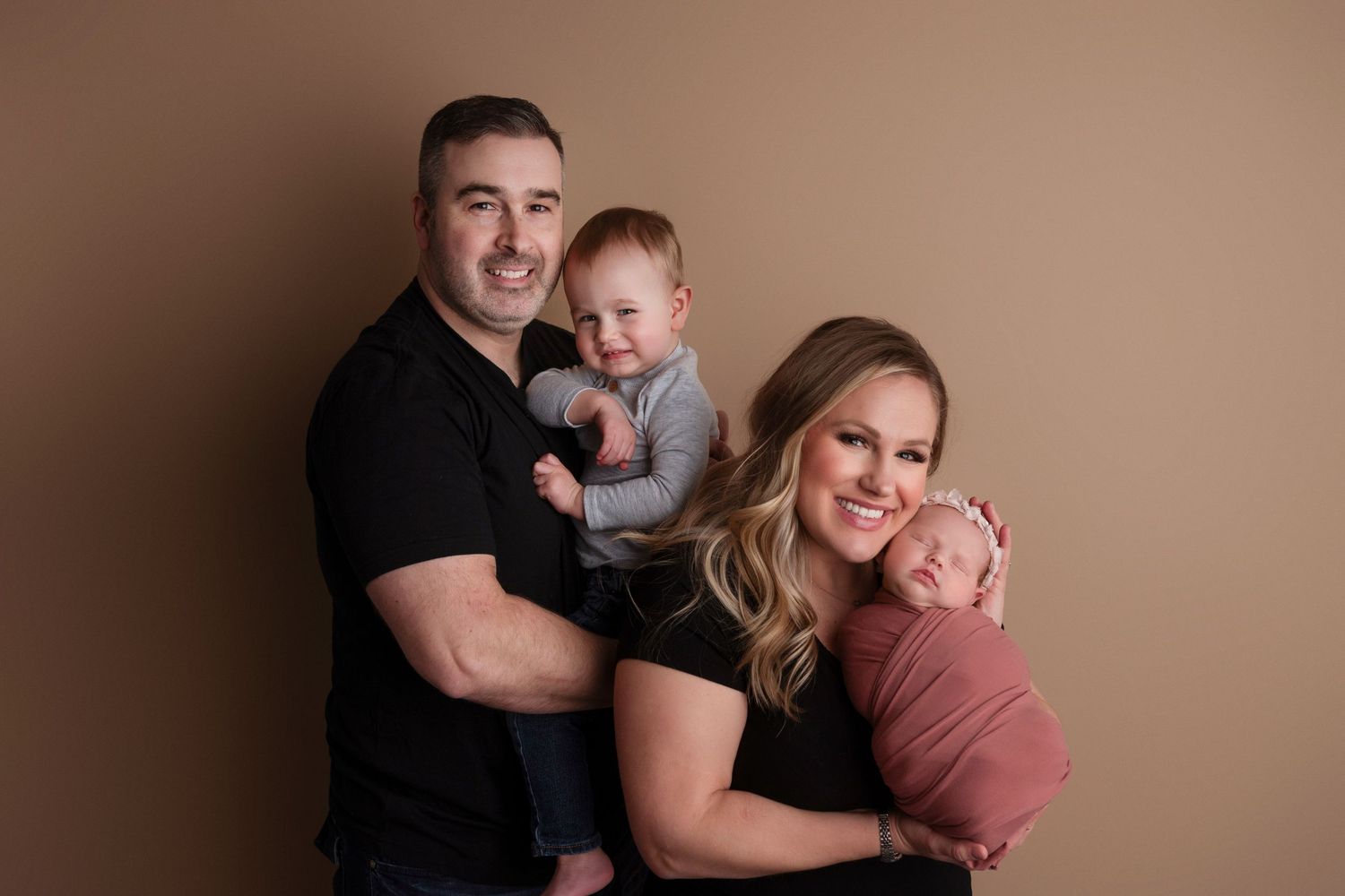 Family portrait against brown backdrop with parents holding two young children in coordinating black and mauve outfits.