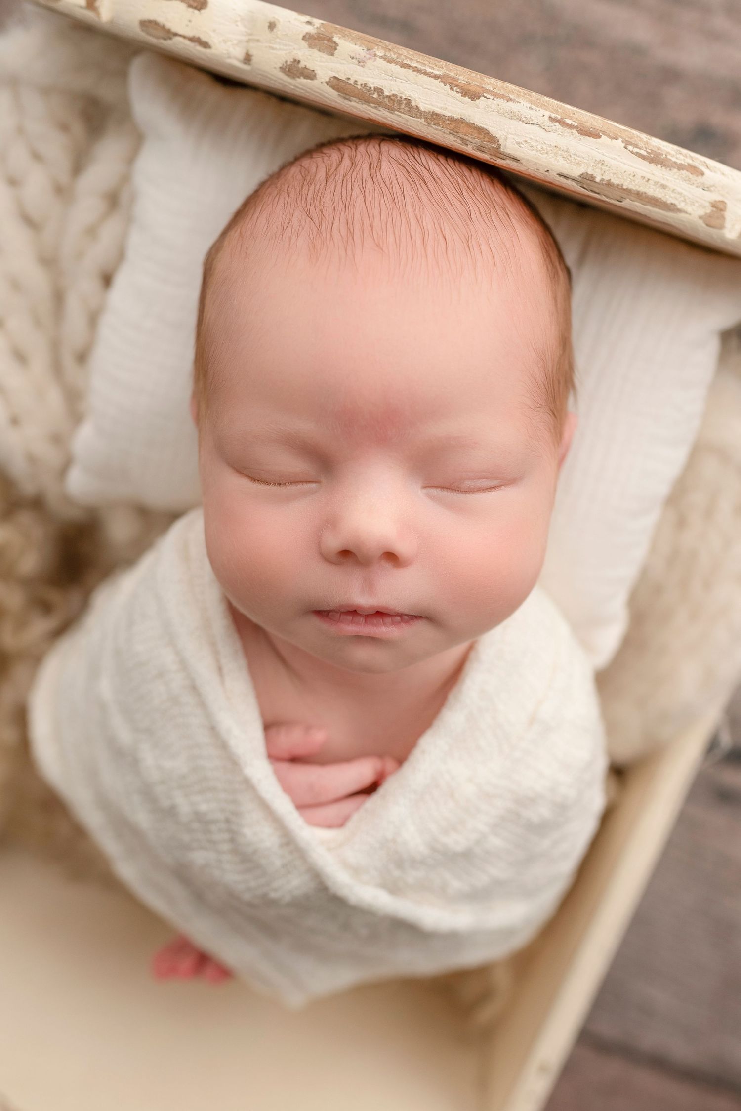 Sleeping newborn baby swaddled in white wrap, wearing a bonnet, resting peacefully in a small wooden bed.