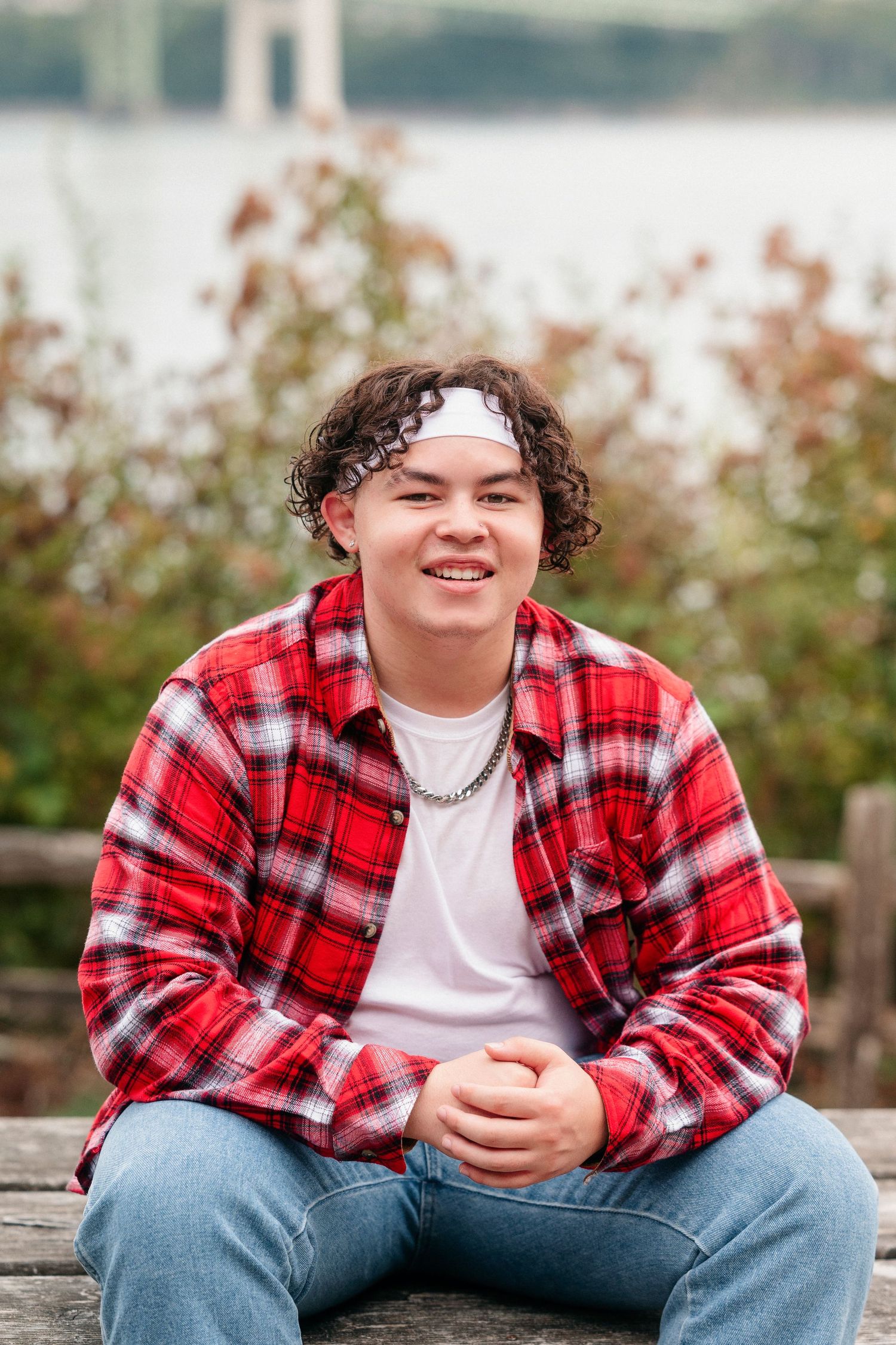 A person in a red plaid flannel shirt and white t-shirt sits casually outdoors with an autumn background.