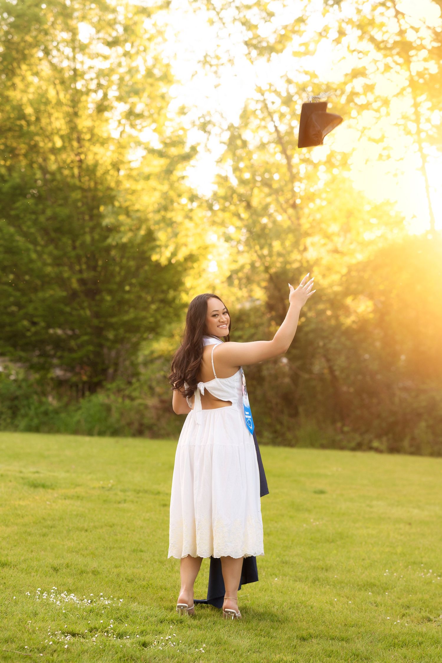 Two graduates in white dresses share a celebratory moment in a grassy field at sunset.