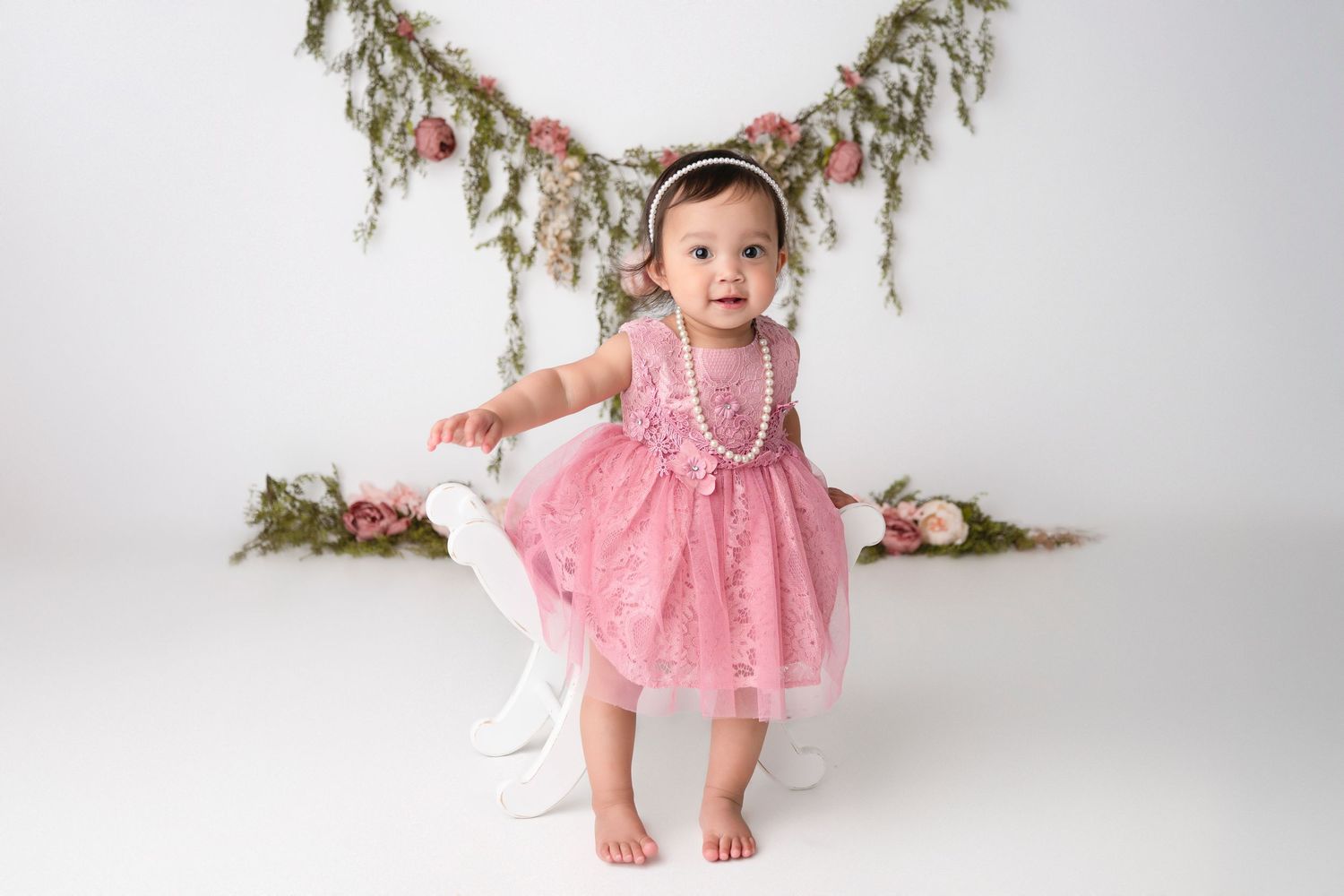Baby wearing pink lace dress and pearls stands against floral garland backdrop in studio setting.