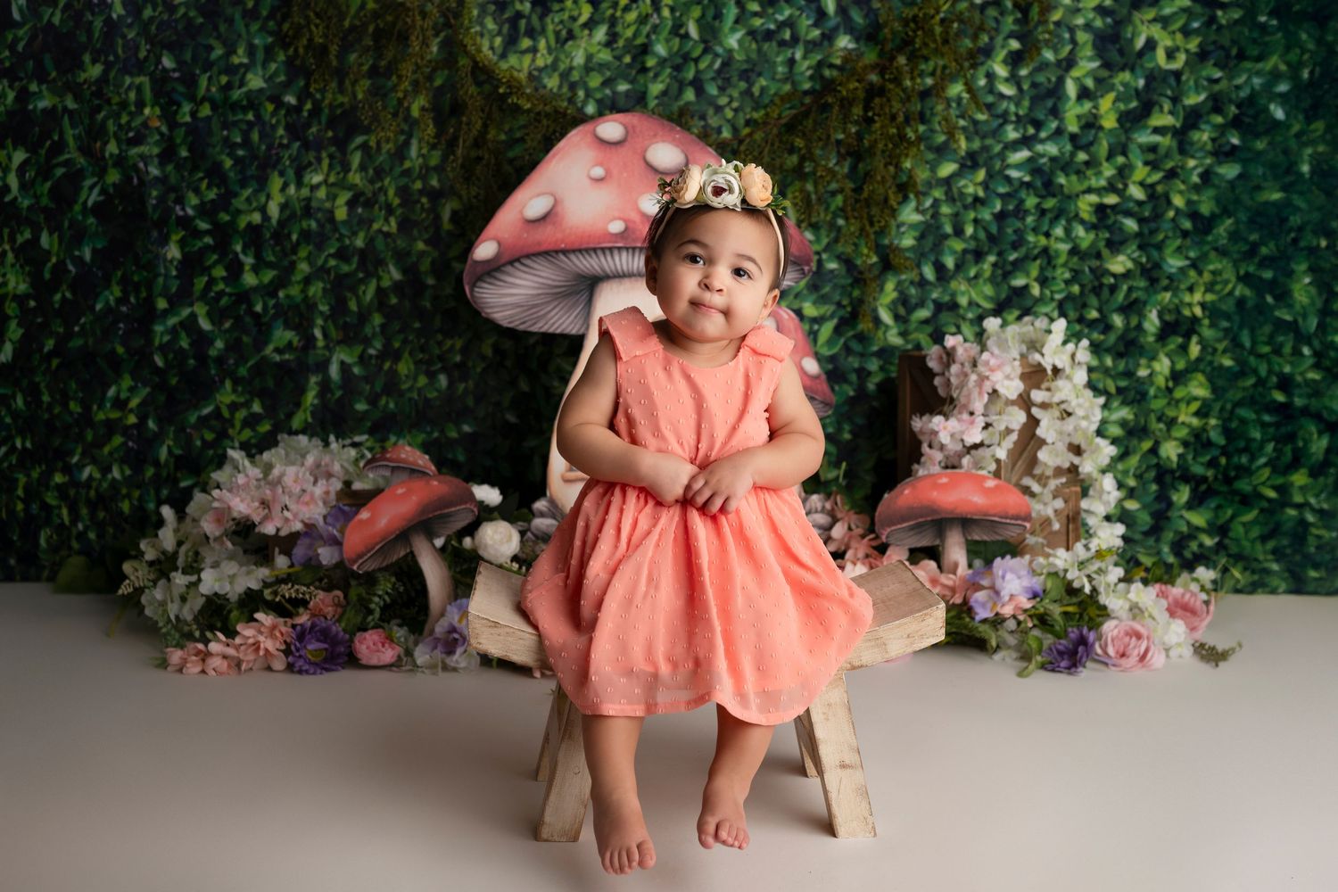 A child in a peach-colored dress sits on a wooden chair against a floral backdrop.