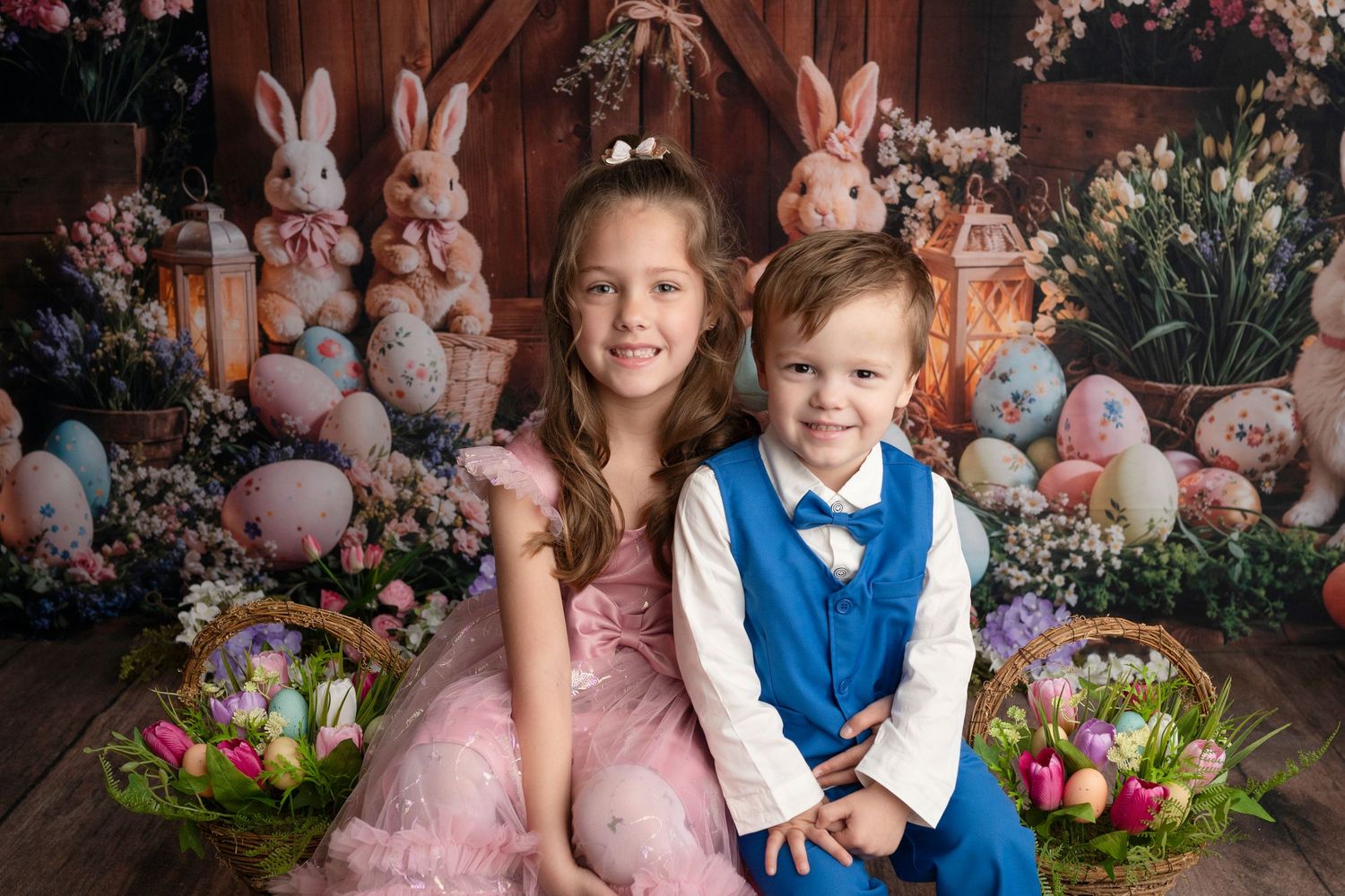 Two children pose for an Easter photo with bunny decorations, colorful eggs, and flower baskets in a festive studio setting.