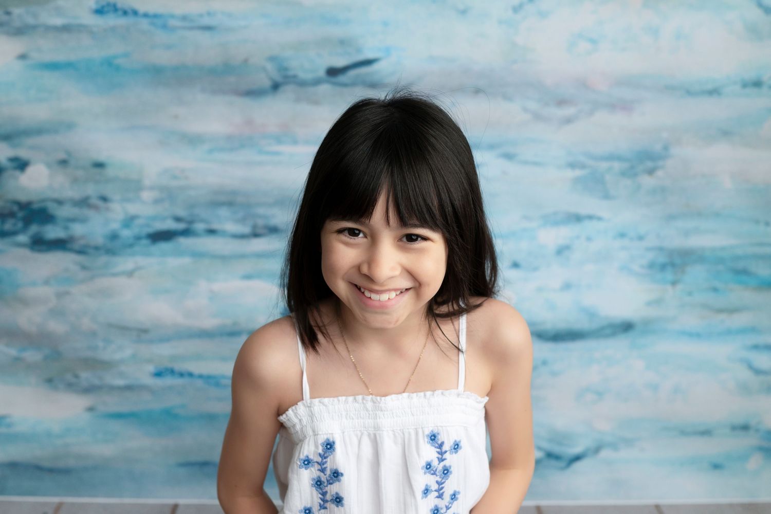 Smiling young girl with dark hair wearing a white floral dress against a blue watercolor backdrop.