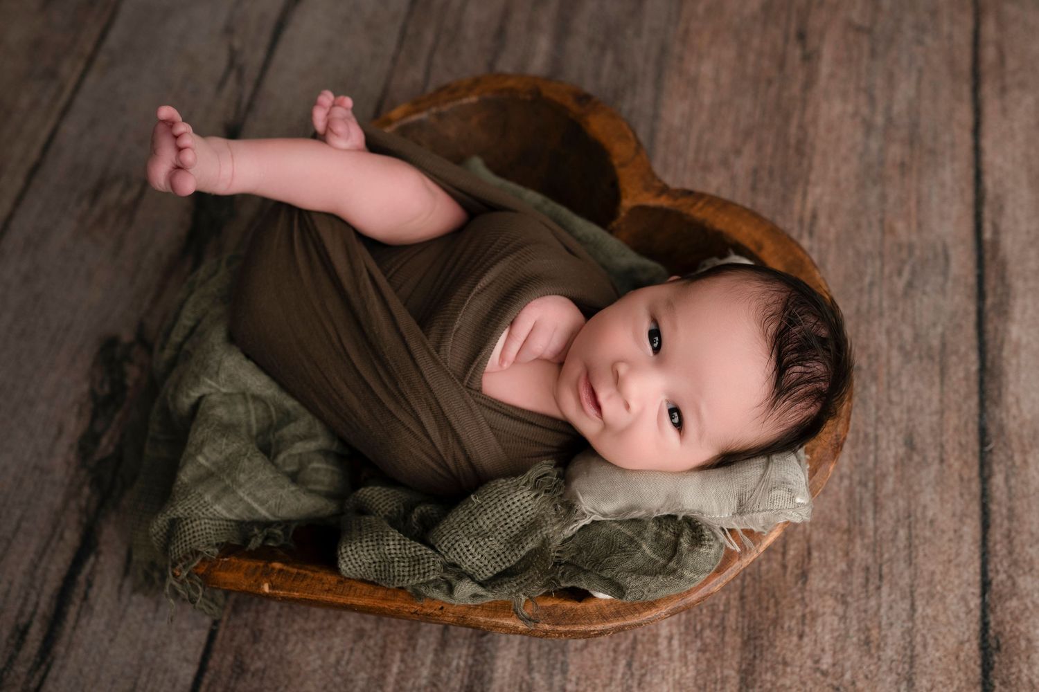 Newborn baby wrapped in olive green swaddle, resting in a wooden heart-shaped bowl on rustic wood floor.