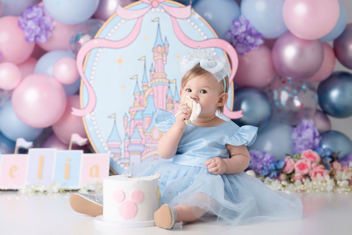 Baby girl in blue dress celebrates first birthday with castle backdrop and colorful balloons at a Disney-themed cake smash.