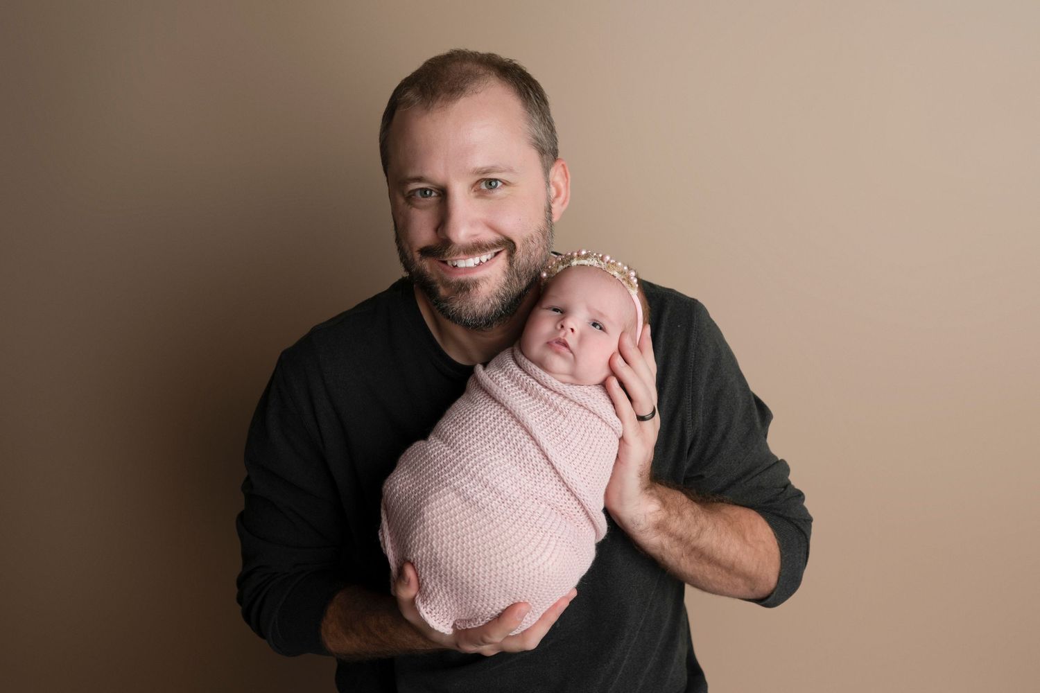 Smiling father holding swaddled newborn baby girl in pink wrap and headband against beige studio backdrop.