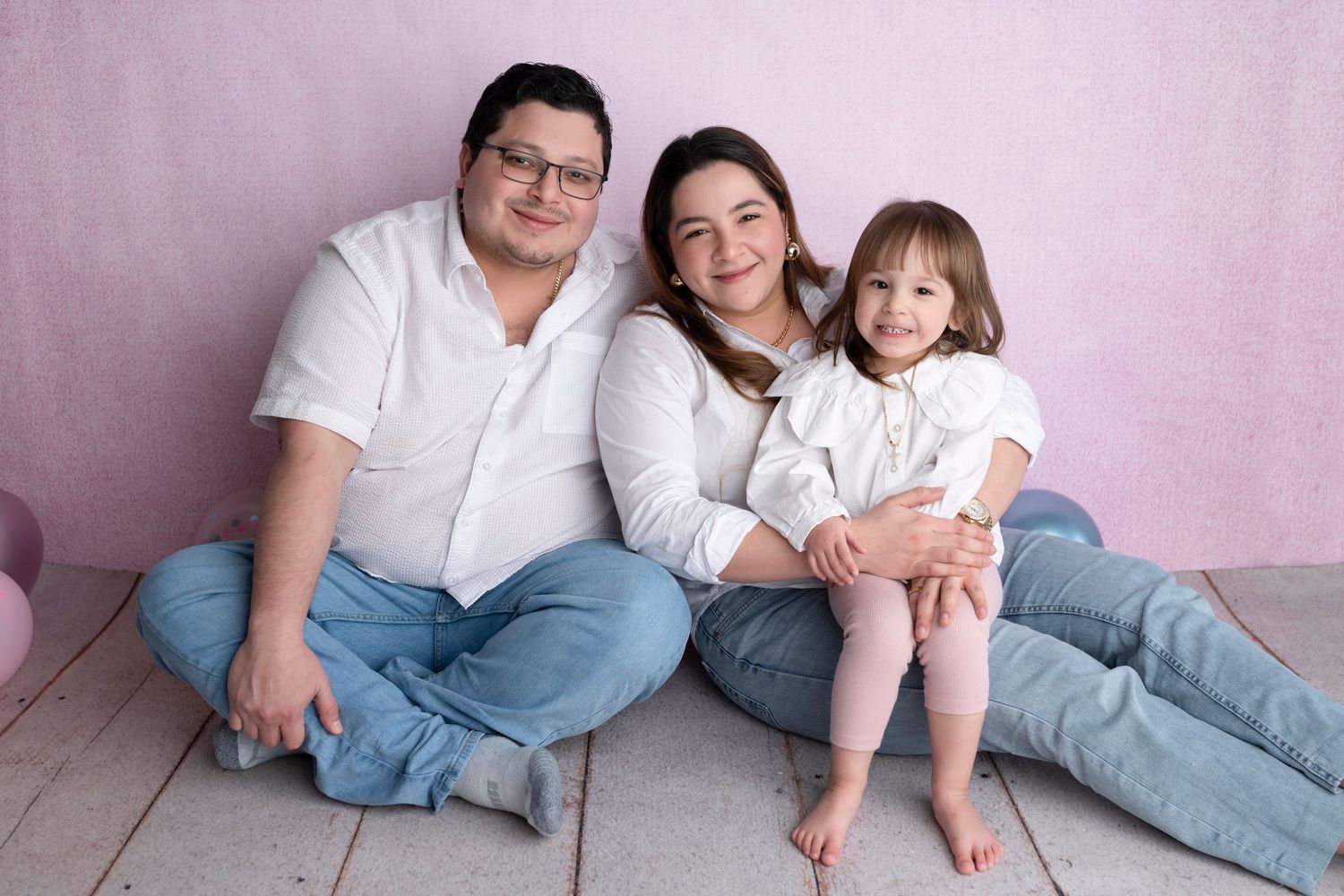 A happy family of three sitting on a wooden floor against a pink background, wearing white shirts and jeans.
