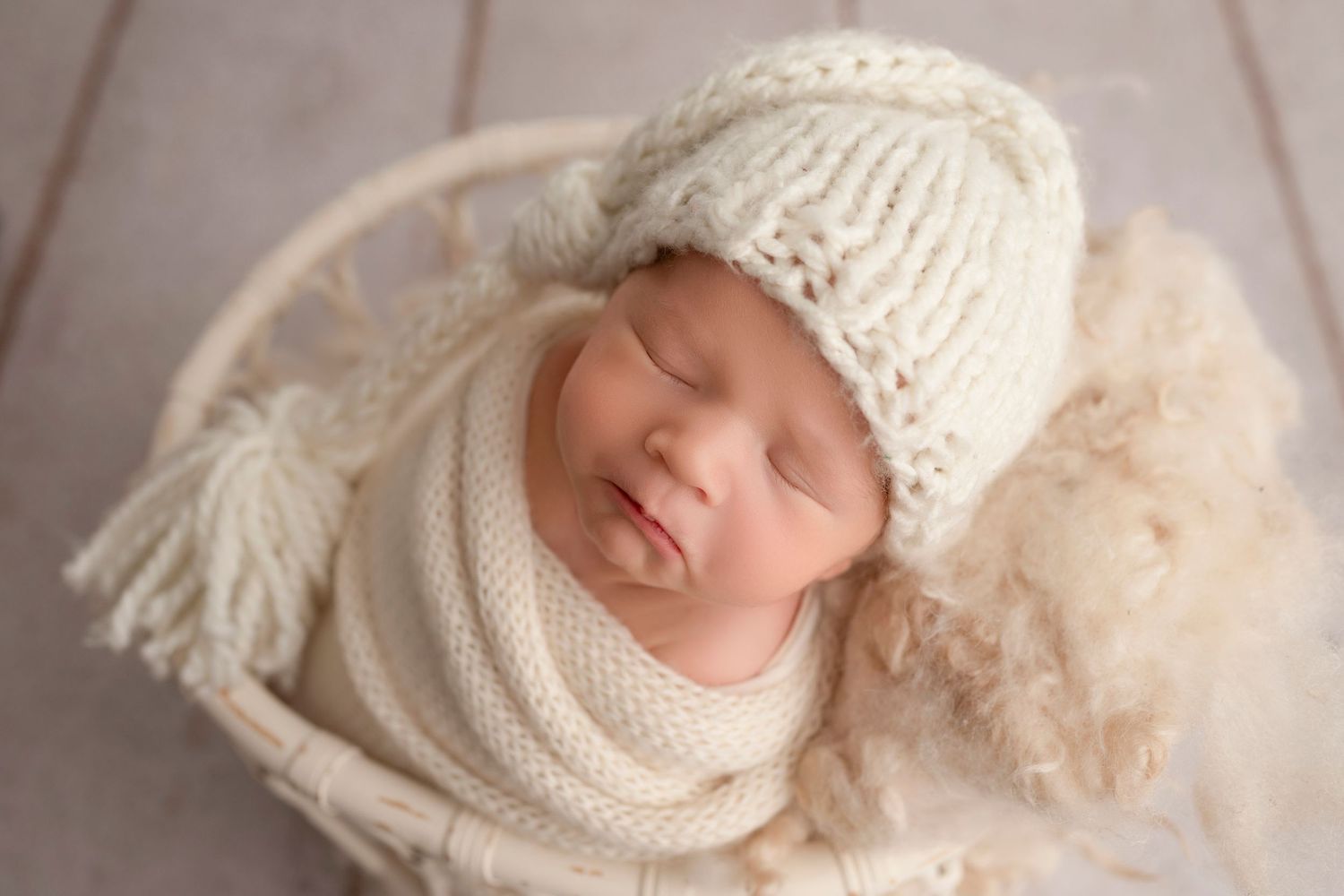 A newborn baby sleeps peacefully wrapped in cozy white knit blankets and wearing a matching hat.