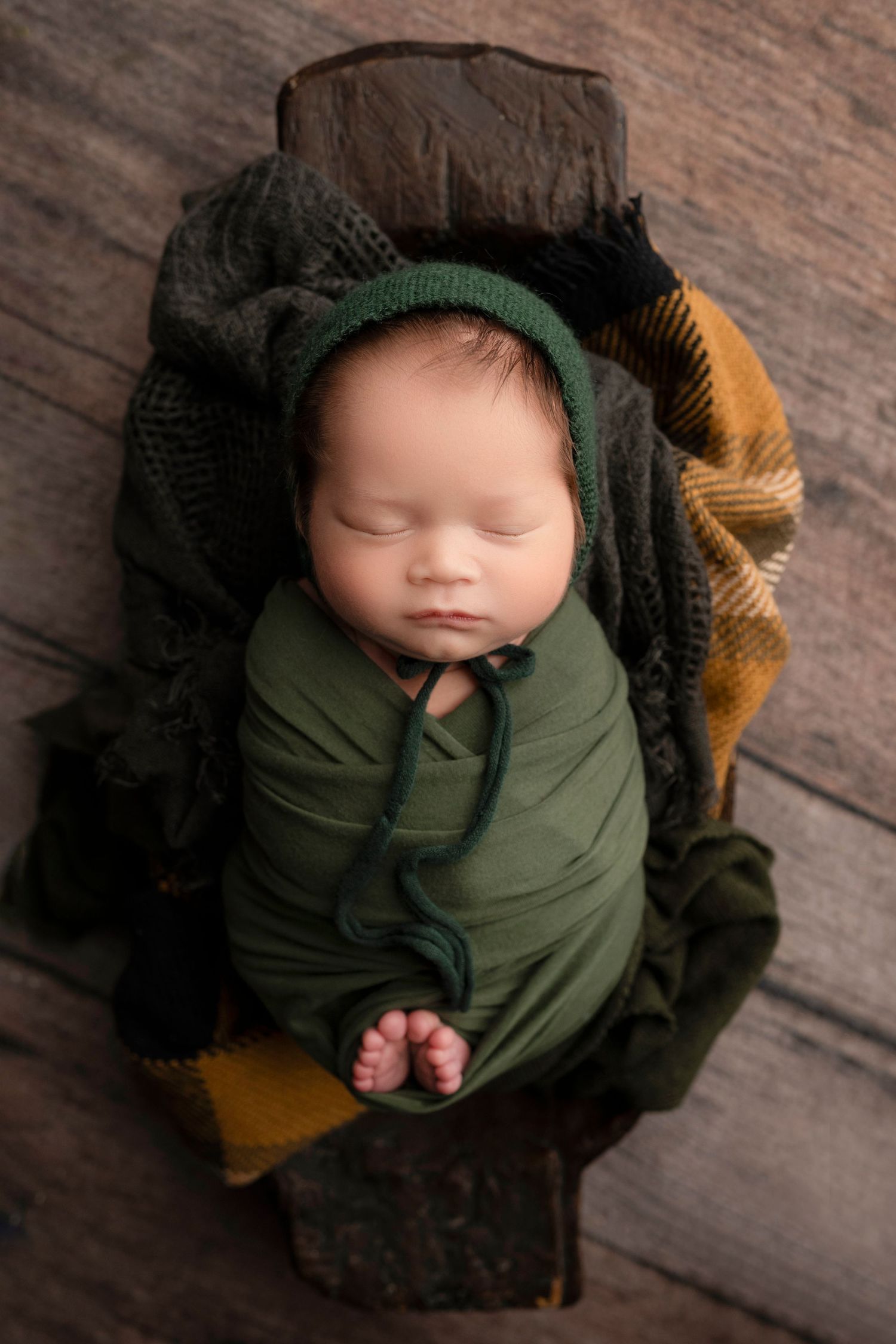 Sleeping newborn wrapped in green and black blankets with a knit cap on wooden floor.