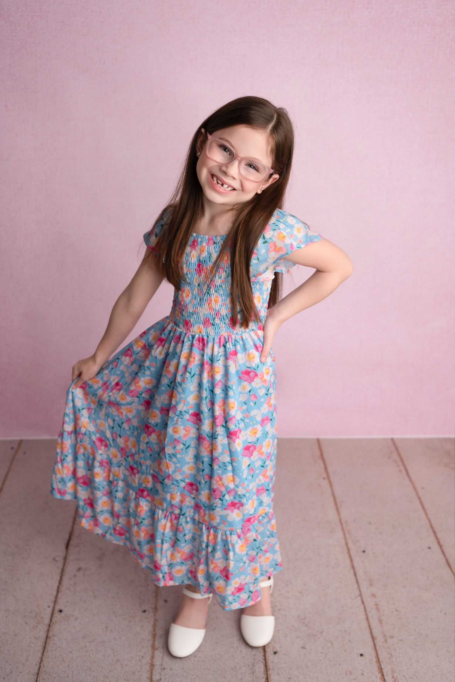 A young child in a floral print dress twirls and poses playfully against a pink backdrop.