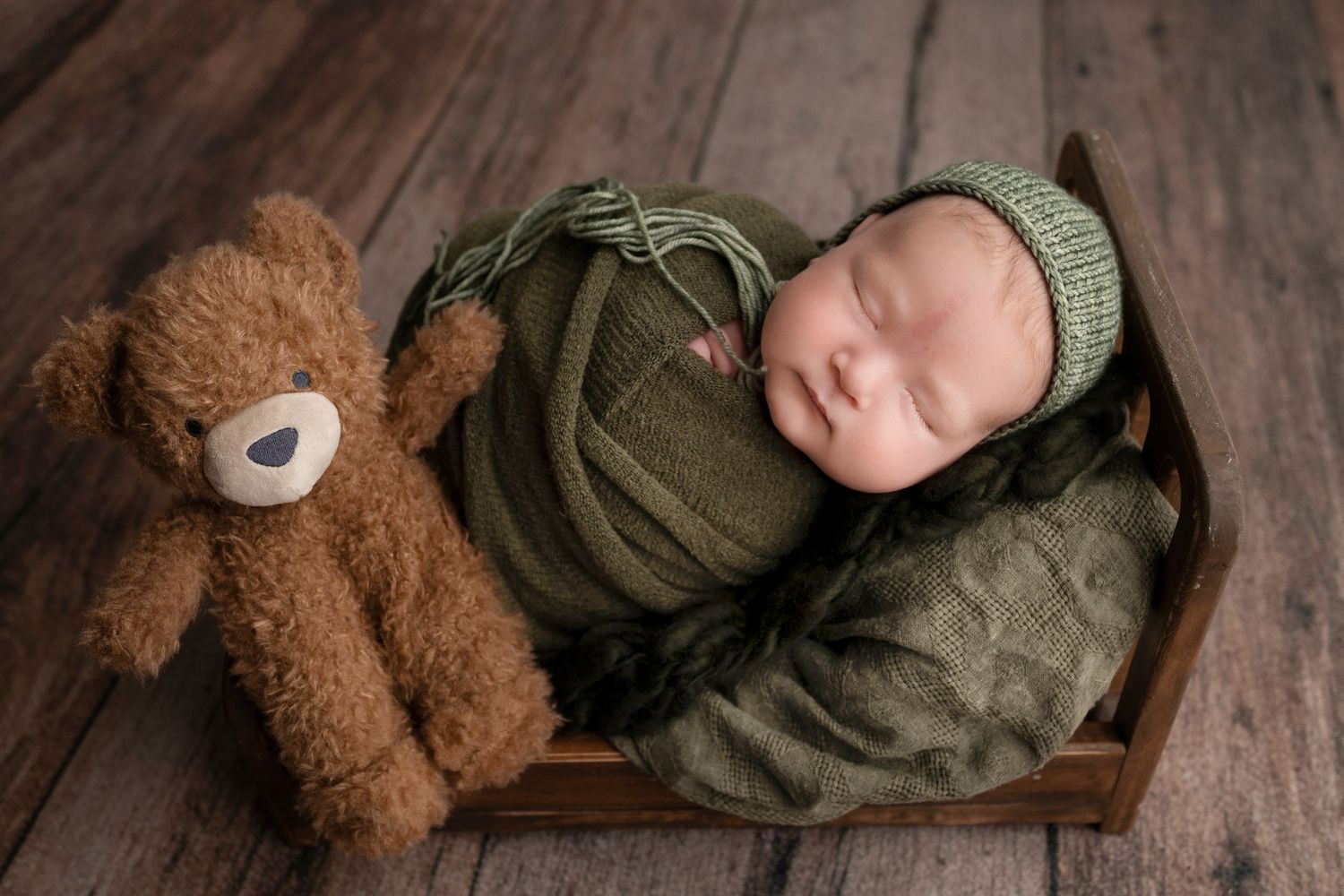 Sleeping newborn baby wrapped in olive green swaddle with knit hat, posed in wooden bed beside brown teddy bear.