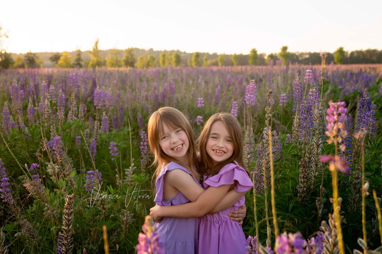 Two smiling girls in purple dresses hugging in a blooming lavender field at golden hour sunset.