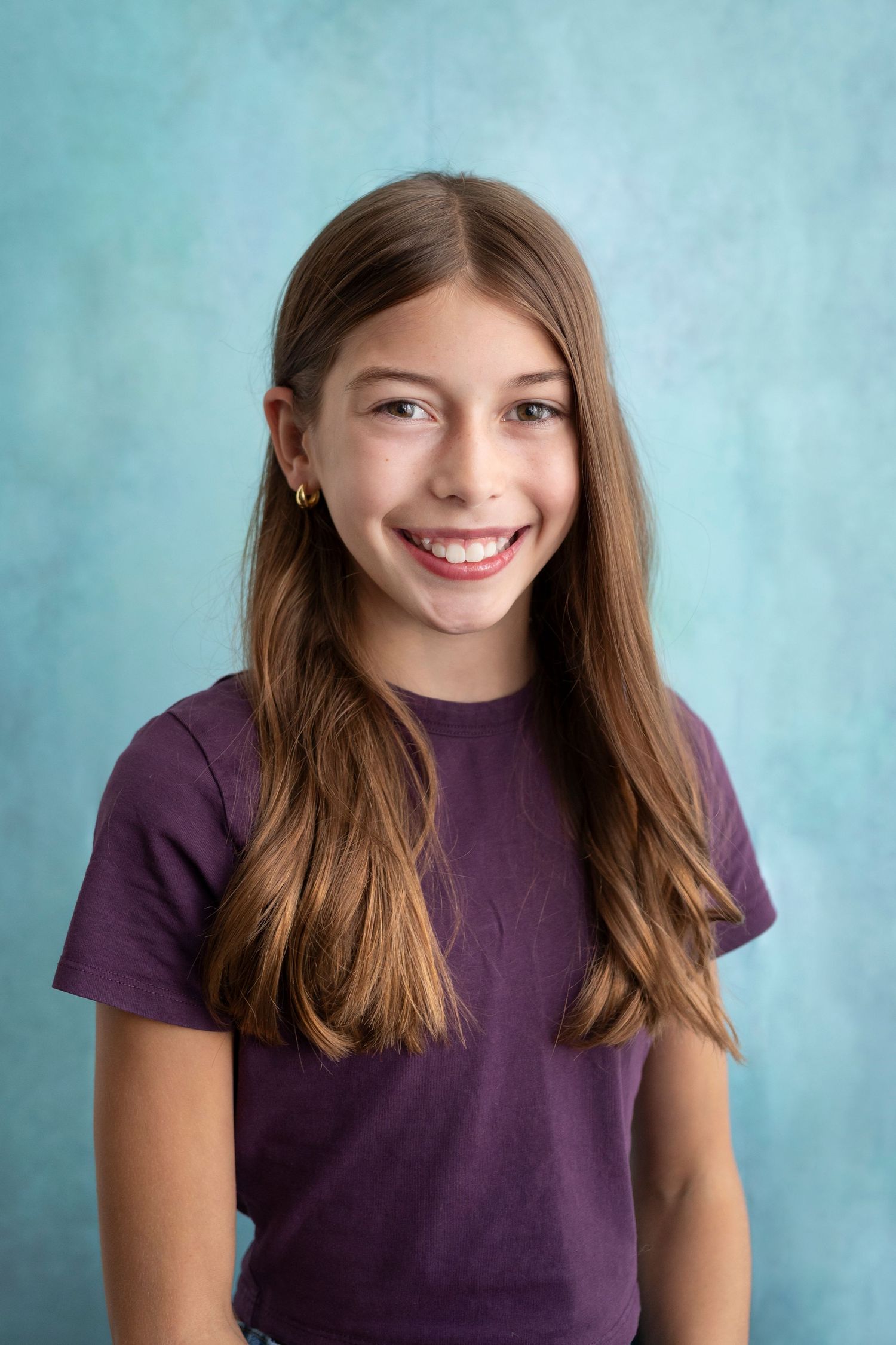 Smiling young girl with long brown hair wearing a purple t-shirt, posed against a blue background.