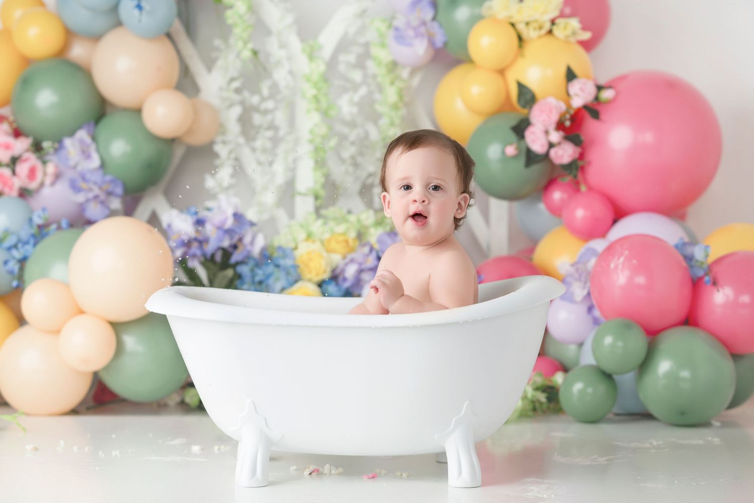 A happy baby sits in a white clawfoot bathtub surrounded by colorful balloons and flowers in a festive photo setup.