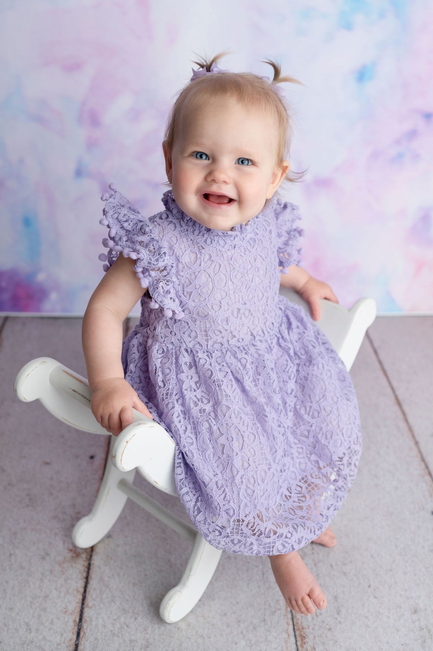 Smiling baby girl in lavender lace dress with pigtails sitting on white chair against pastel backdrop.