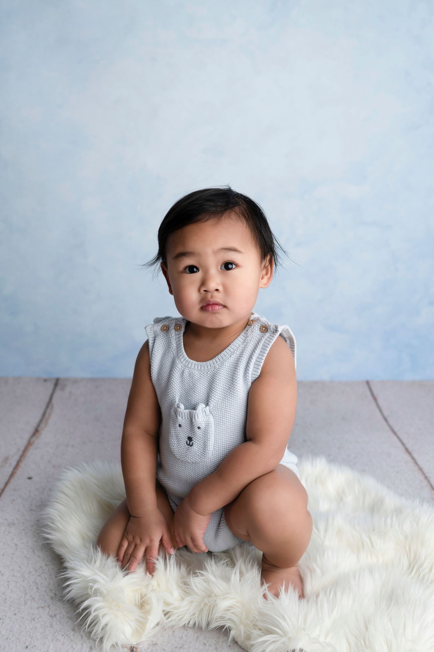 A cute Asian baby in a light blue knit romper sits on a fluffy white rug against a soft blue backdrop.