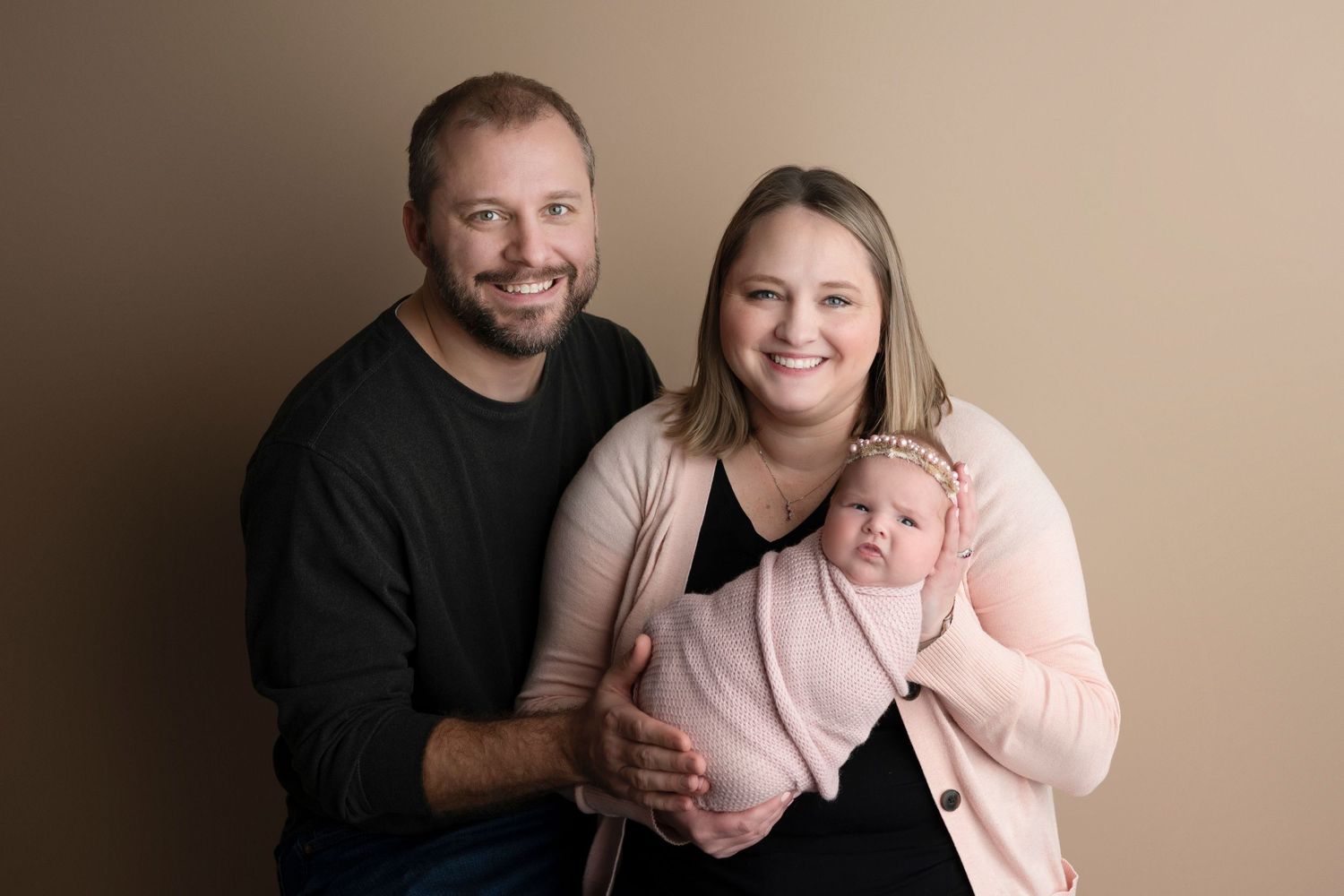 Smiling couple holding their newborn baby wrapped in pink during a professional studio portrait session.