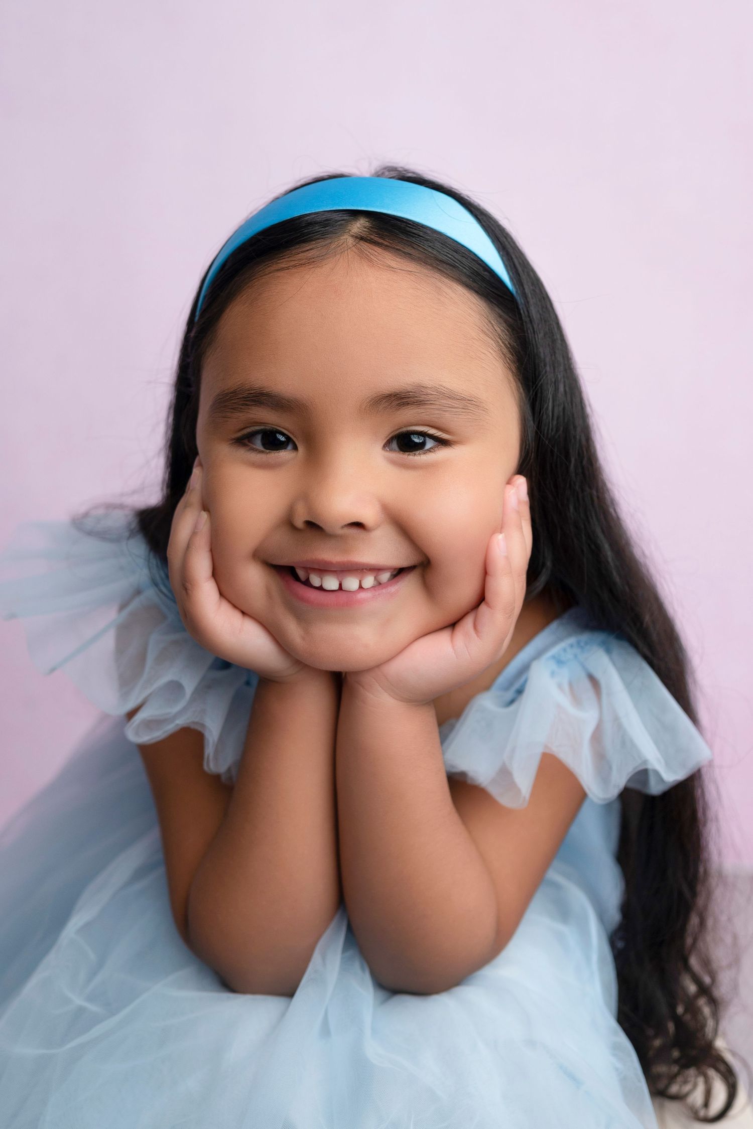 A smiling young girl in a blue tulle dress and headband poses with hands on cheeks against a pink background.