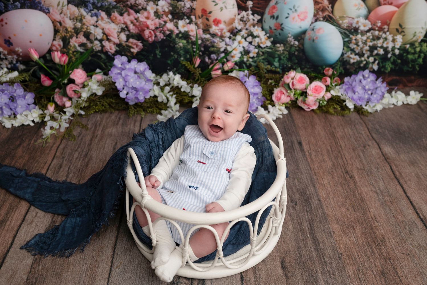 Smiling baby in white outfit sitting in wicker basket surrounded by colorful Easter eggs and flowers on wood floor.
