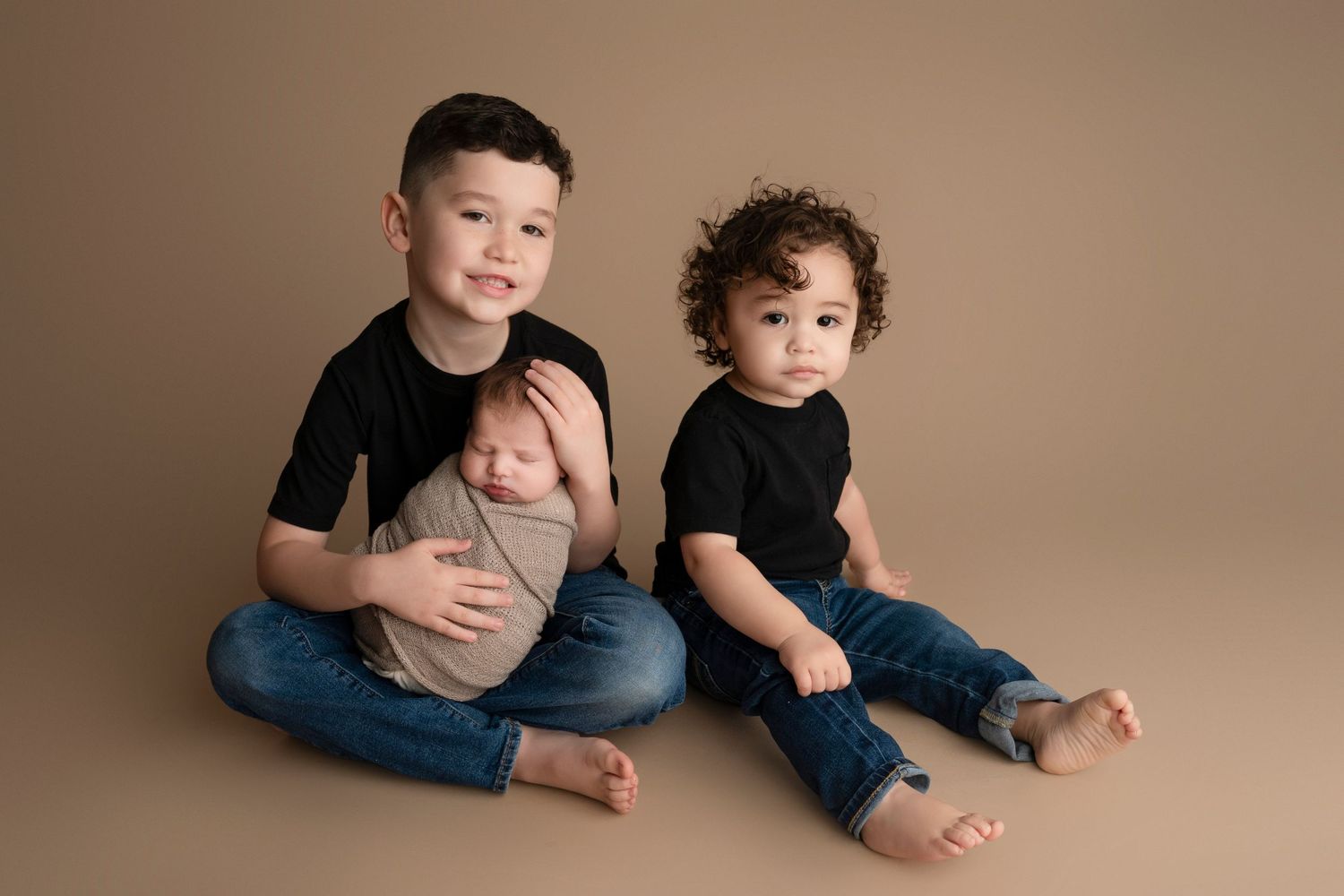 Two older brothers sit together on a tan backdrop, gently holding their sleeping newborn sibling in a professional studio portrait.