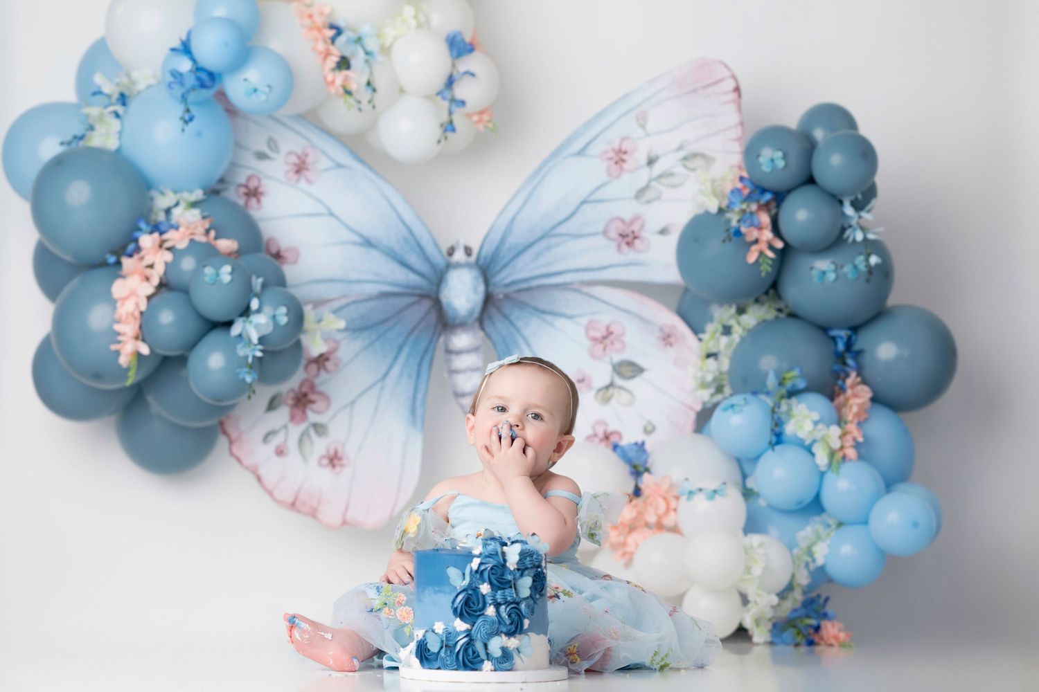 Whimsical first birthday setup with blue balloon arch, butterflies, and soft pastel decorations.