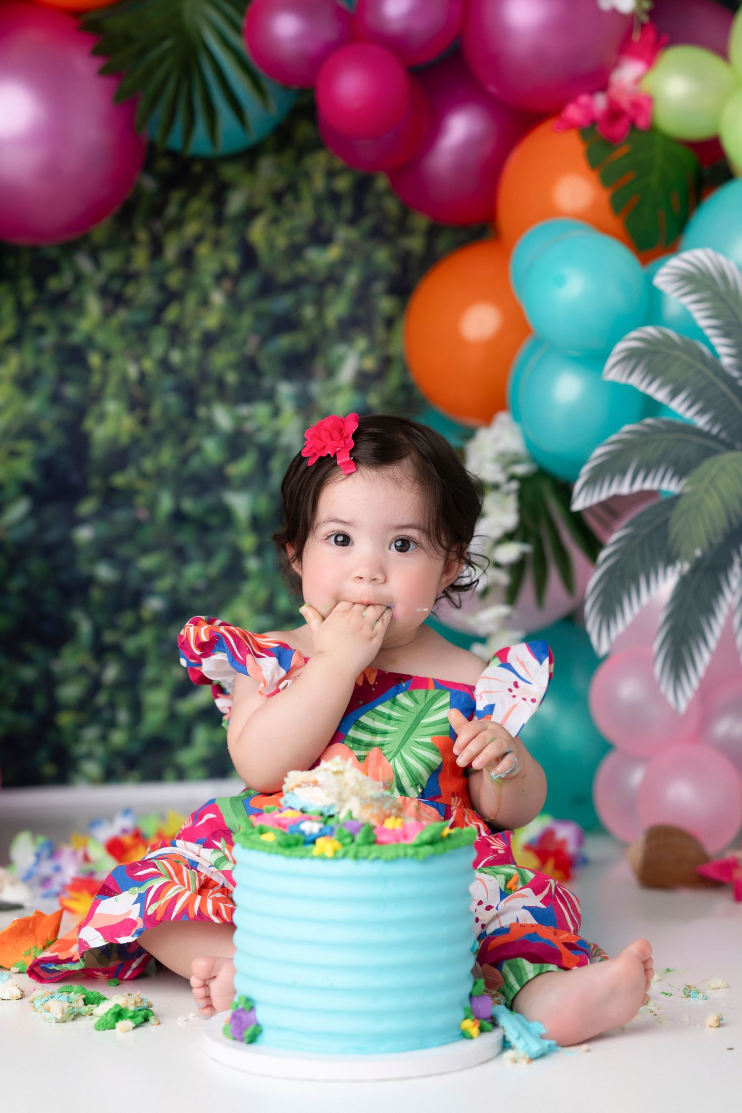 Colorful balloon arch and tropical decor create a festive cake smash photo session with bright teal and pink colors.