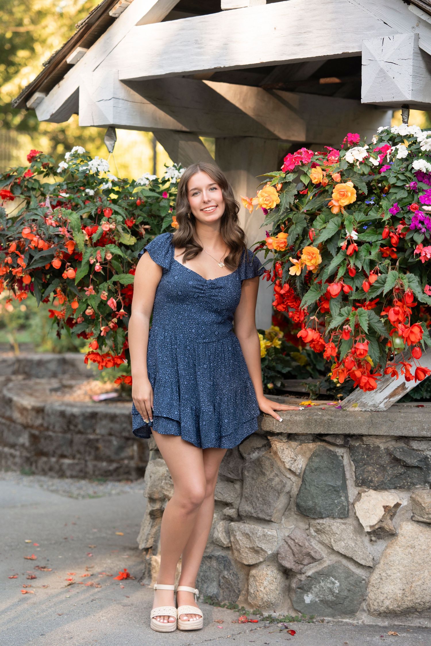 A person in a blue dress poses near a stone wall with vibrant red and orange flowers in the background.