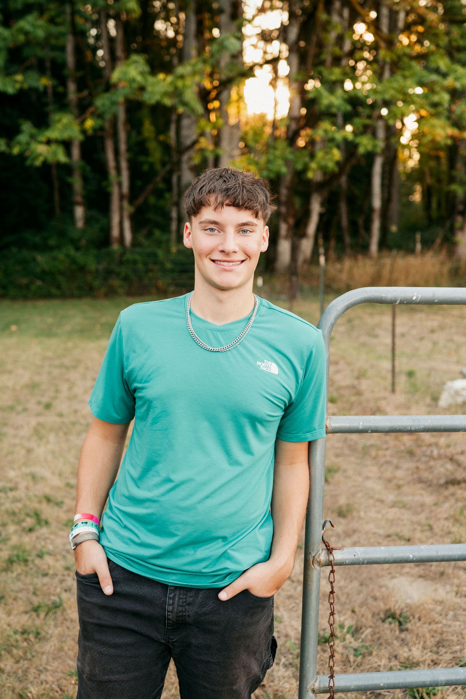 A person in a teal t-shirt and dark jeans poses outdoors against a wooded background during golden hour.