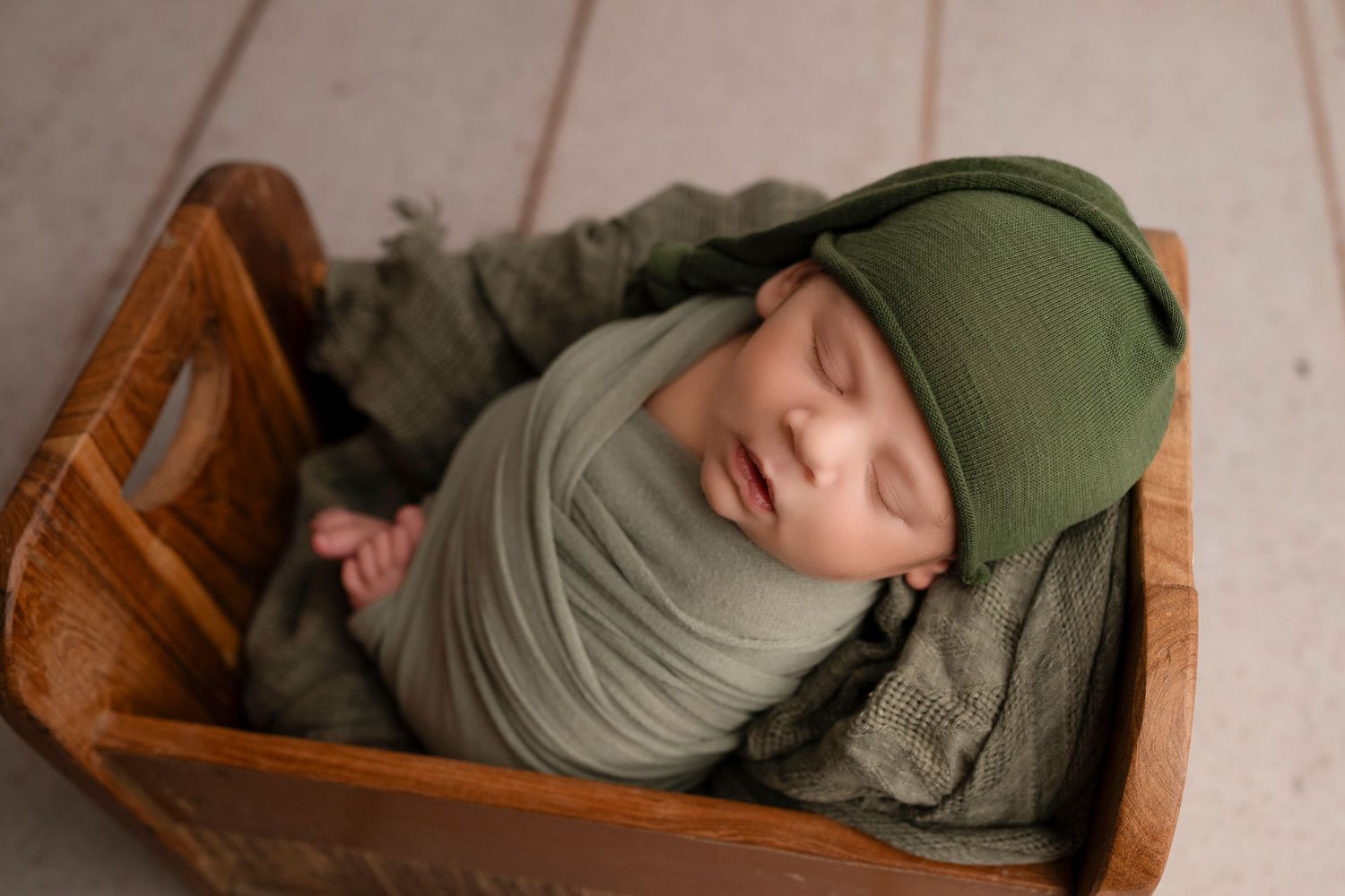 A sleeping newborn wrapped in olive green swaddle lies peacefully in a wooden bed.