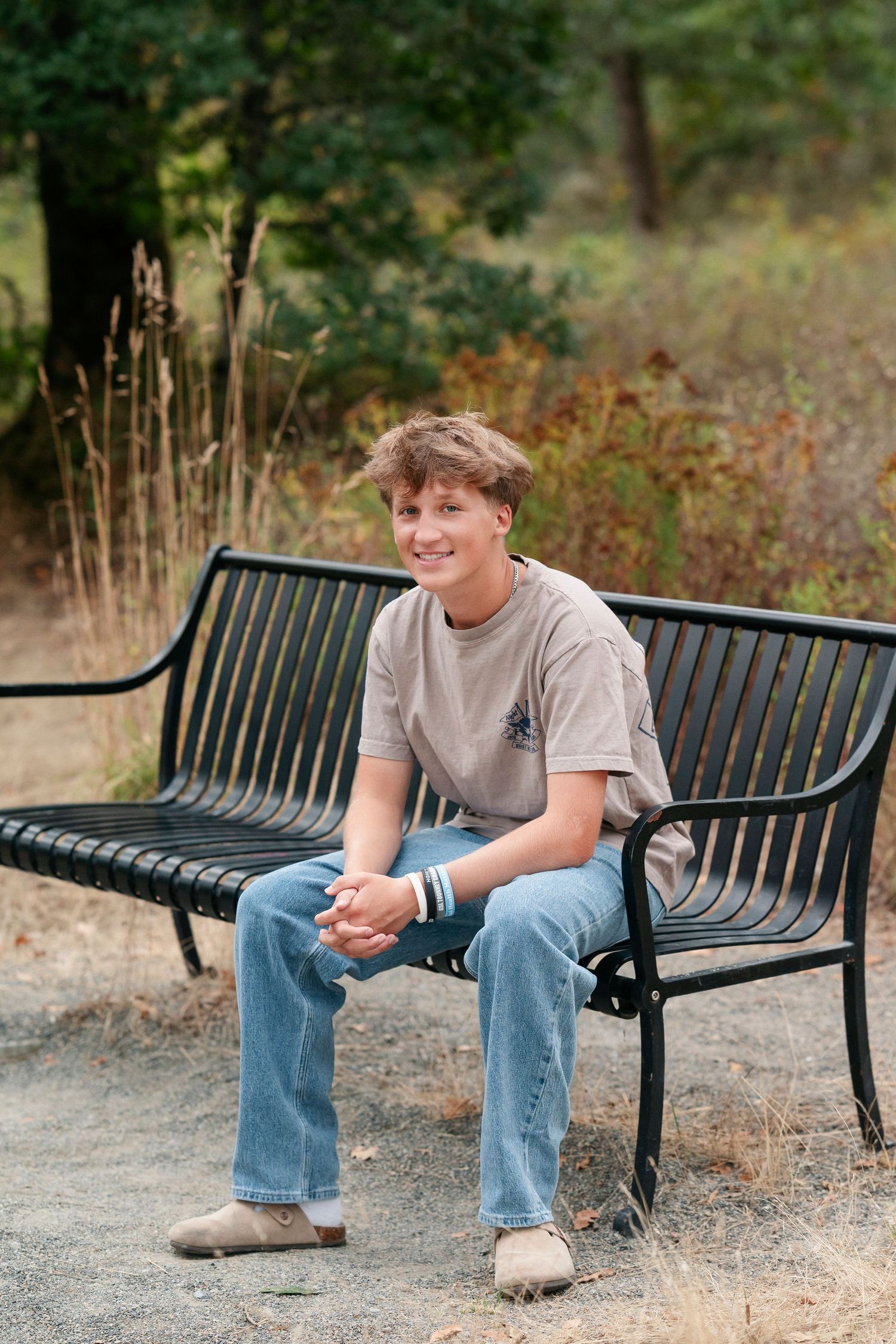 A person in jeans and a gray t-shirt sits casually on a black metal park bench in an outdoor setting.