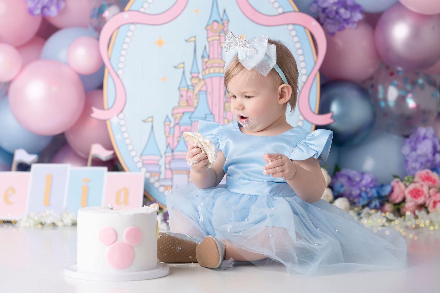 Baby girl in blue dress at Disney-themed first birthday cake smash with pink and blue balloons and castle backdrop.