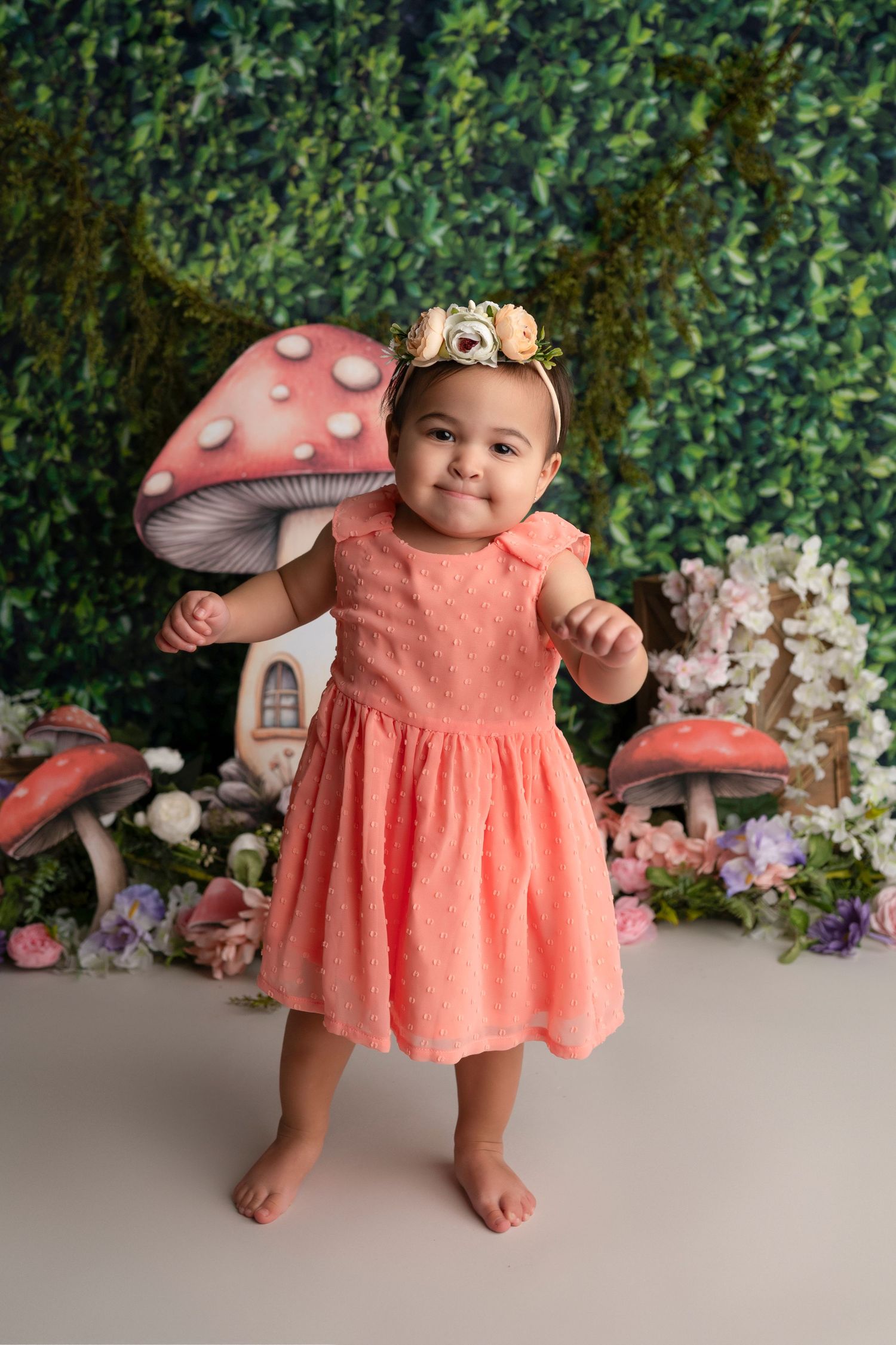 A child in a coral dress poses against a green backdrop with decorative mushrooms and floral elements in a studio setting.