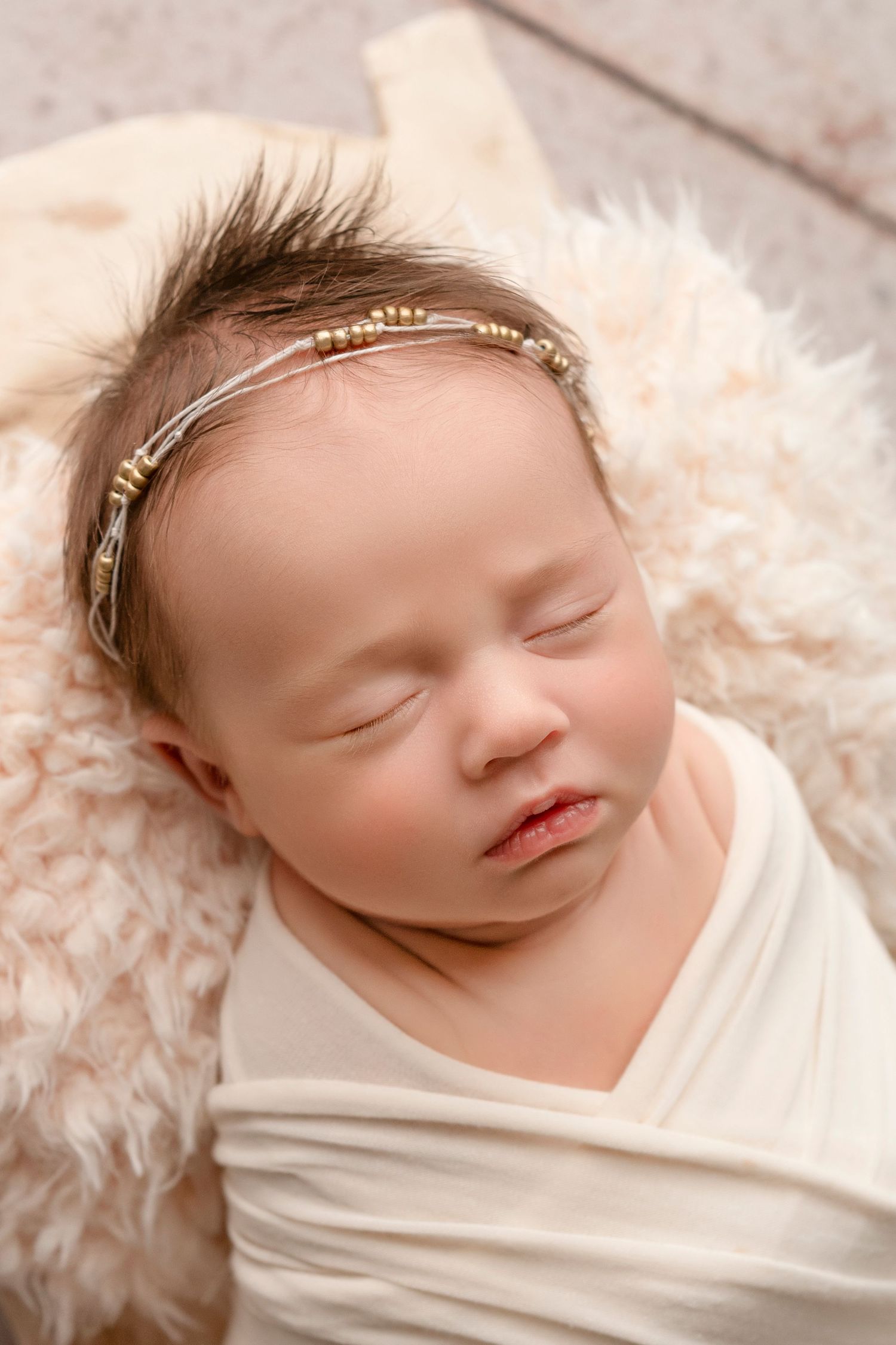 Sleeping newborn wrapped in white fabric wearing delicate headband rests peacefully on soft furry blanket.