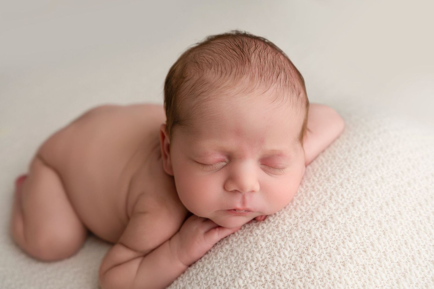 Peaceful sleeping newborn baby curled up on soft white textured blanket during professional photo session.