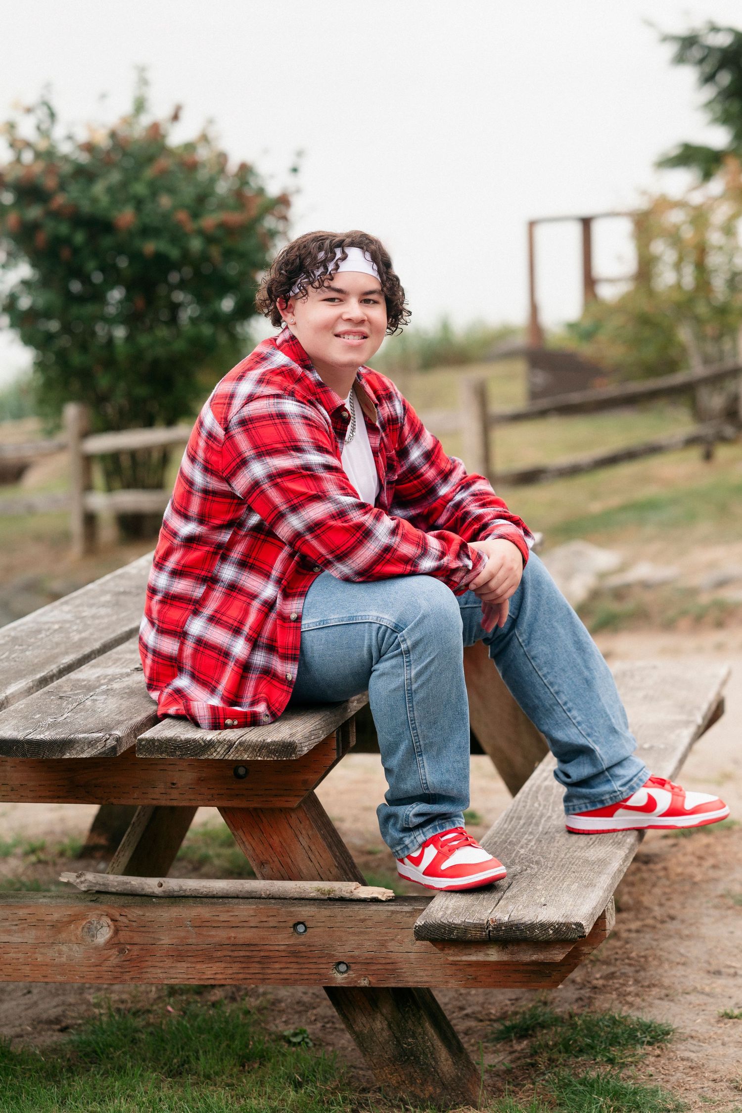 A person in red plaid and jeans sits on a wooden picnic table with red sneakers and a natural backdrop.