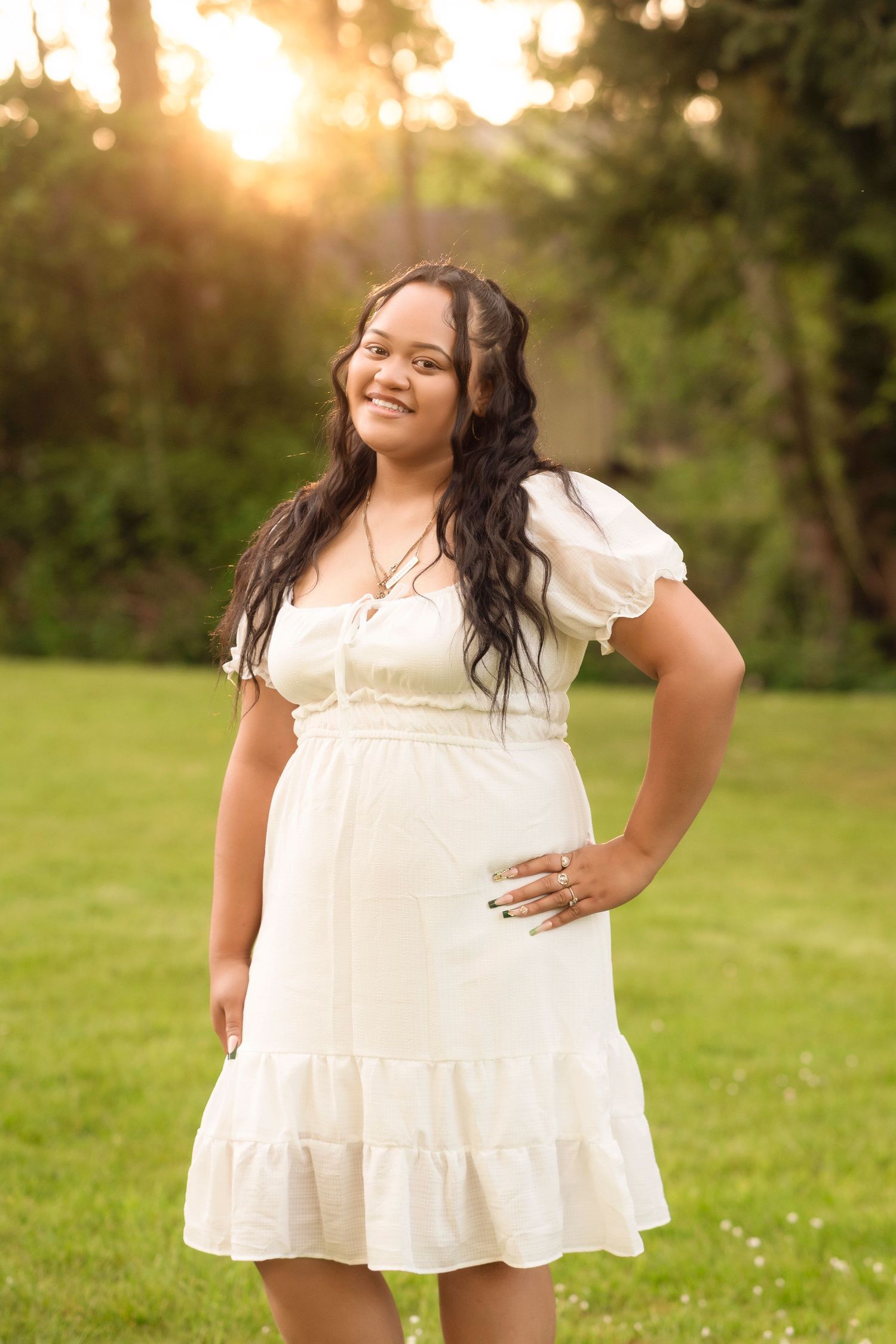 Person in flowing white summer dress poses outdoors during golden hour in a grassy field.