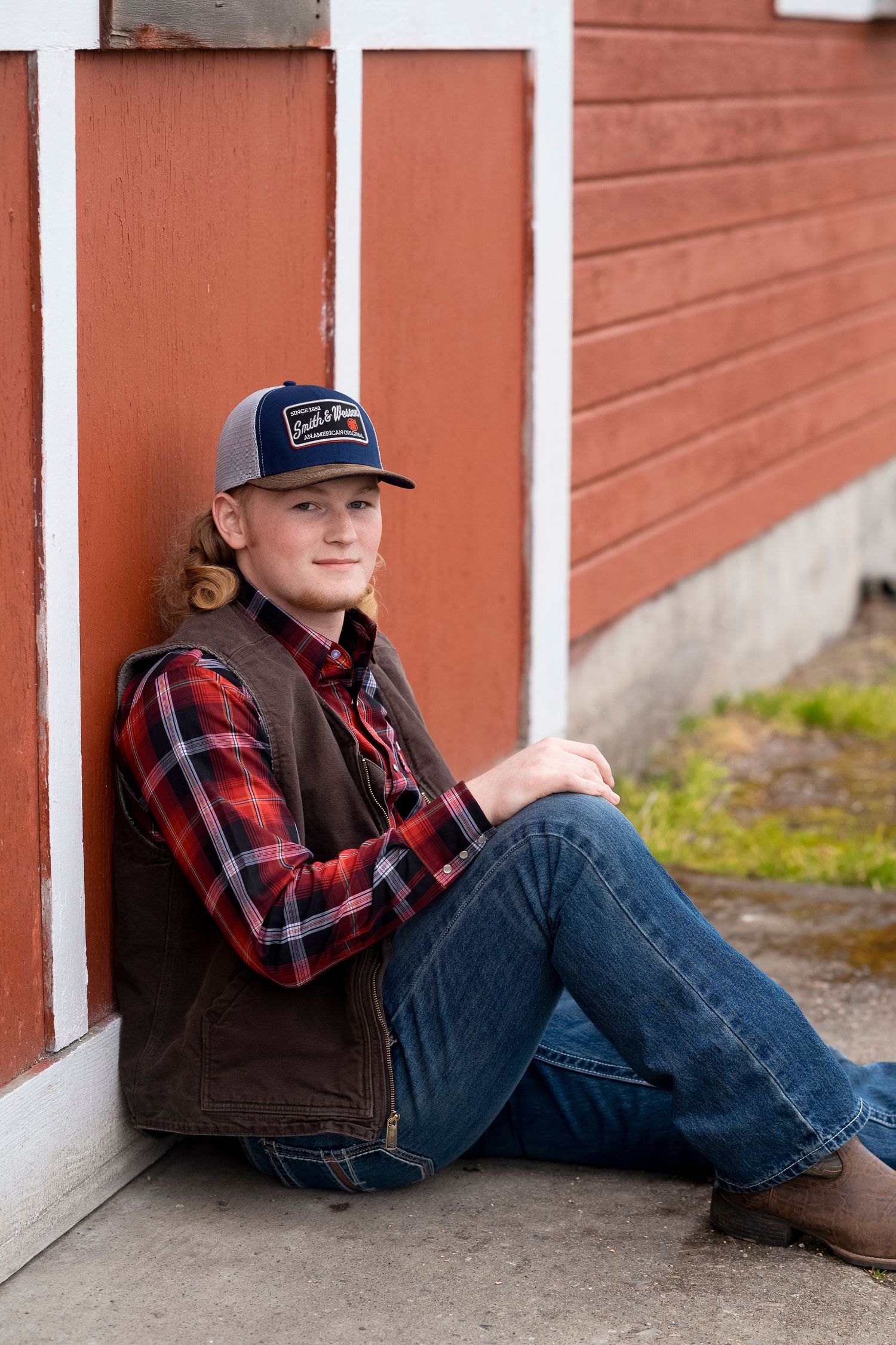 Young person in plaid shirt and vest sits casually against red barn wall wearing baseball cap and jeans.