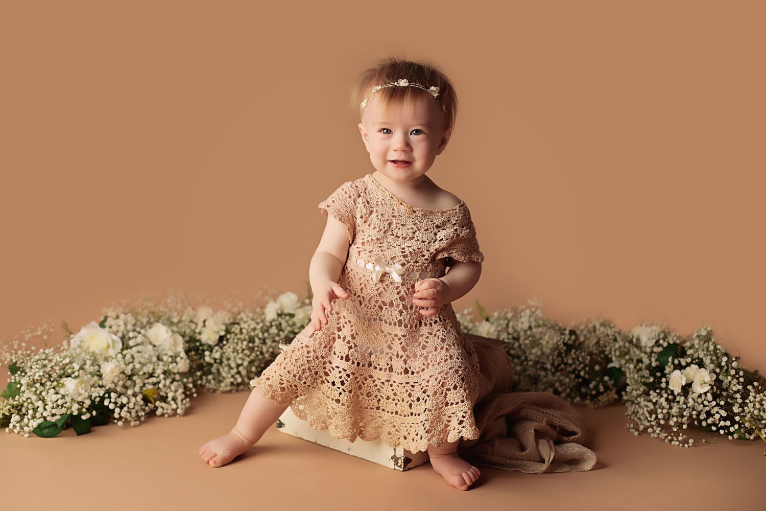 A small child in a champagne lace dress sits on a brown backdrop surrounded by delicate white flowers.