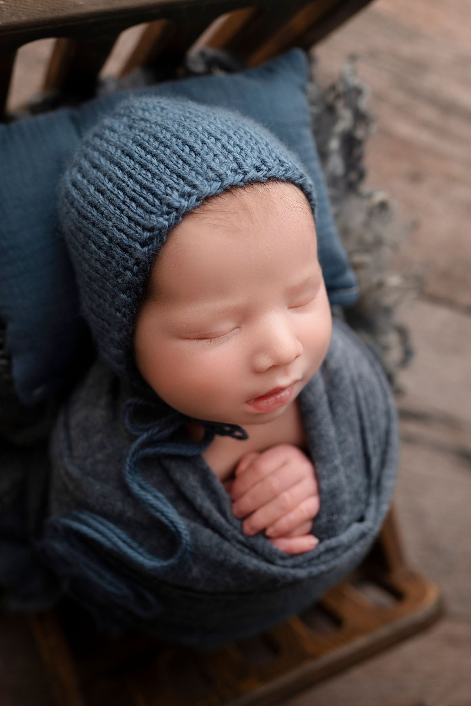Sleeping newborn baby wrapped in blue knit fabric wearing a matching bonnet, posed in a wooden prop.
