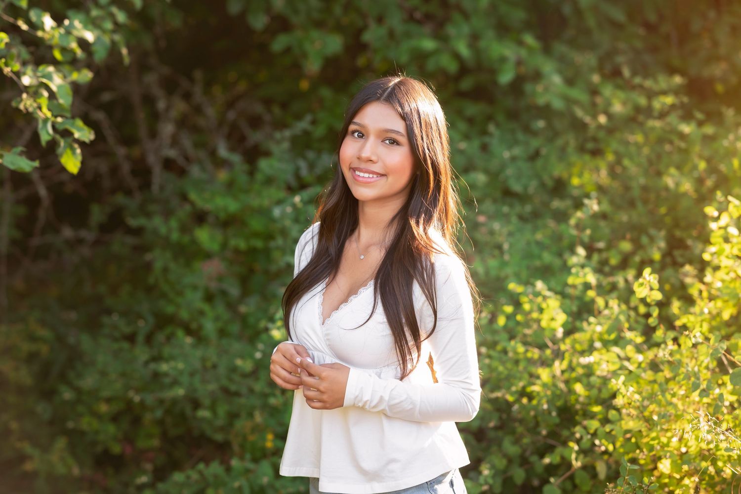 Young woman in white blazer smiling outdoors, surrounded by lush green foliage in warm sunlight.